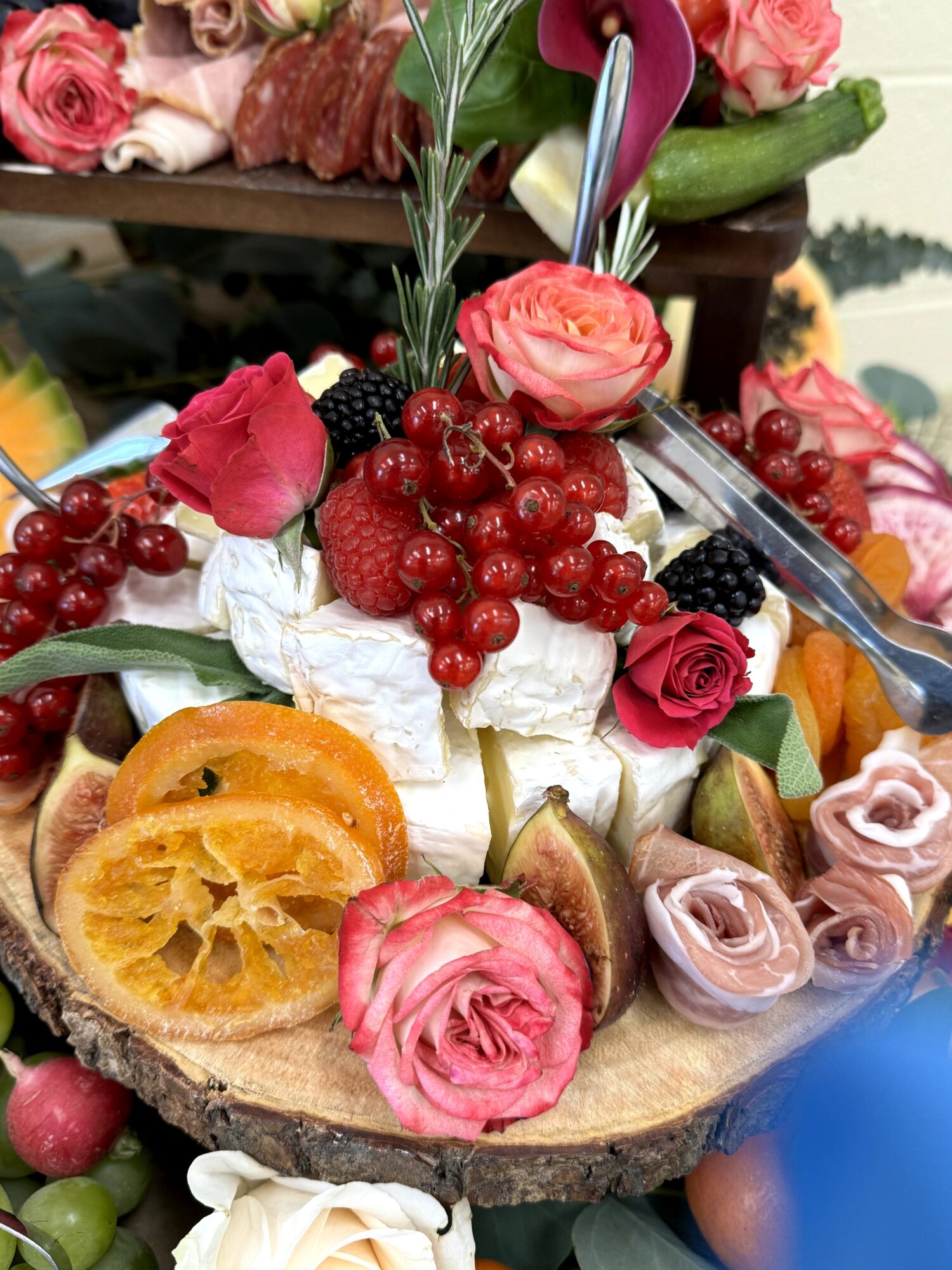 Decorative cake with roses, berries, and fruit slices on a wooden base, surrounded by flowers and greenery.