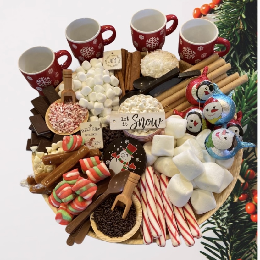 Festive holiday tray with red mugs, marshmallows, candies, cinnamon sticks, and snow-themed decorations.
