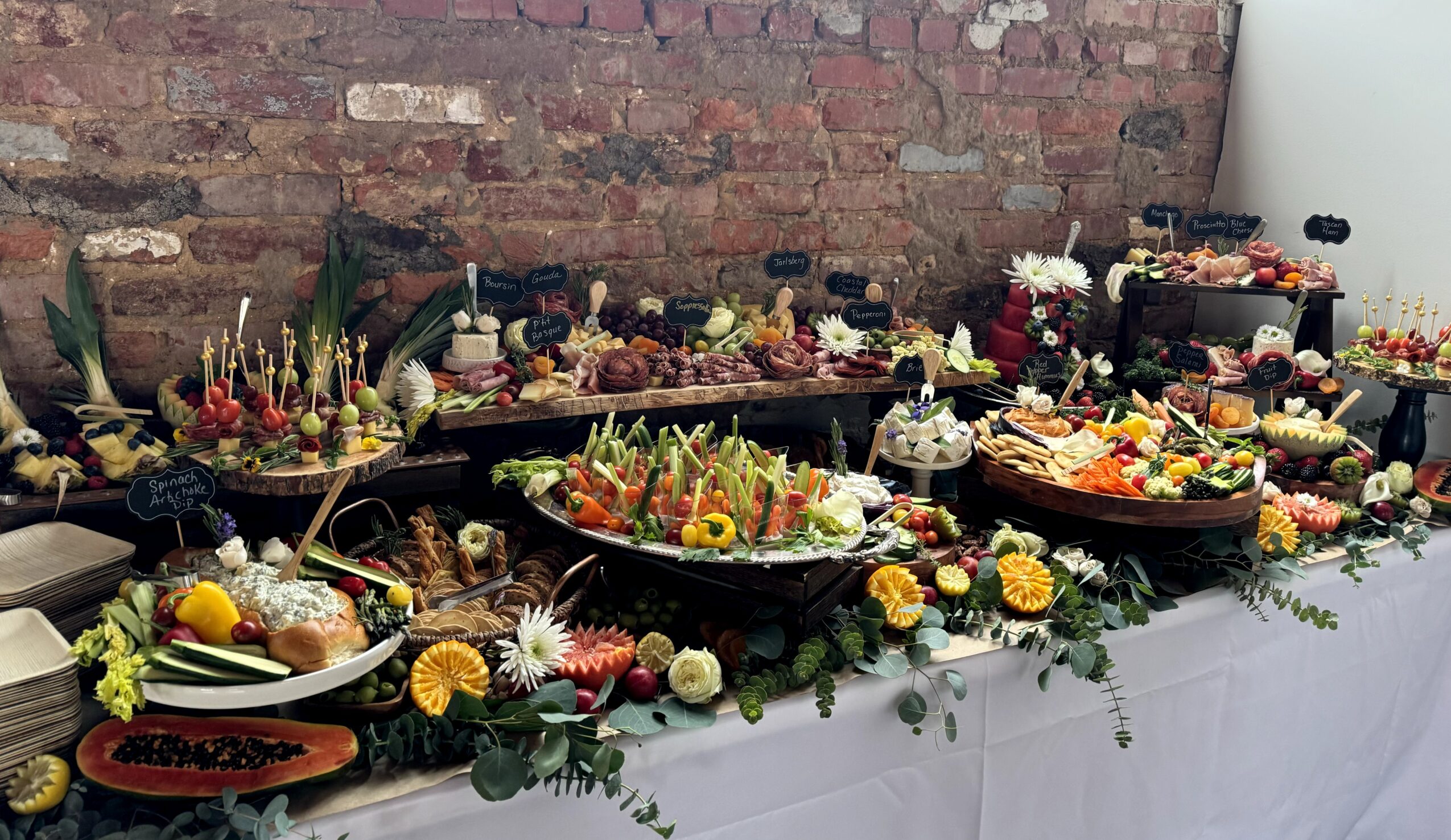 Table with various colorful food dishes and floral decorations against a brick wall, arranged for a celebration or event.