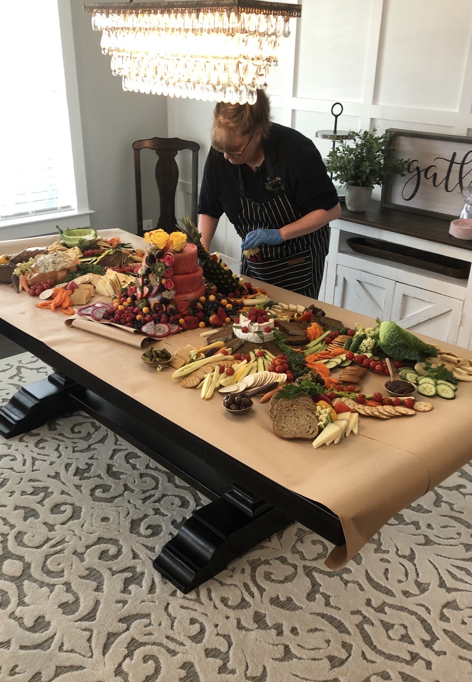 Person arranging a large charcuterie board with various cheeses, fruits, and vegetables on a table in a bright kitchen.