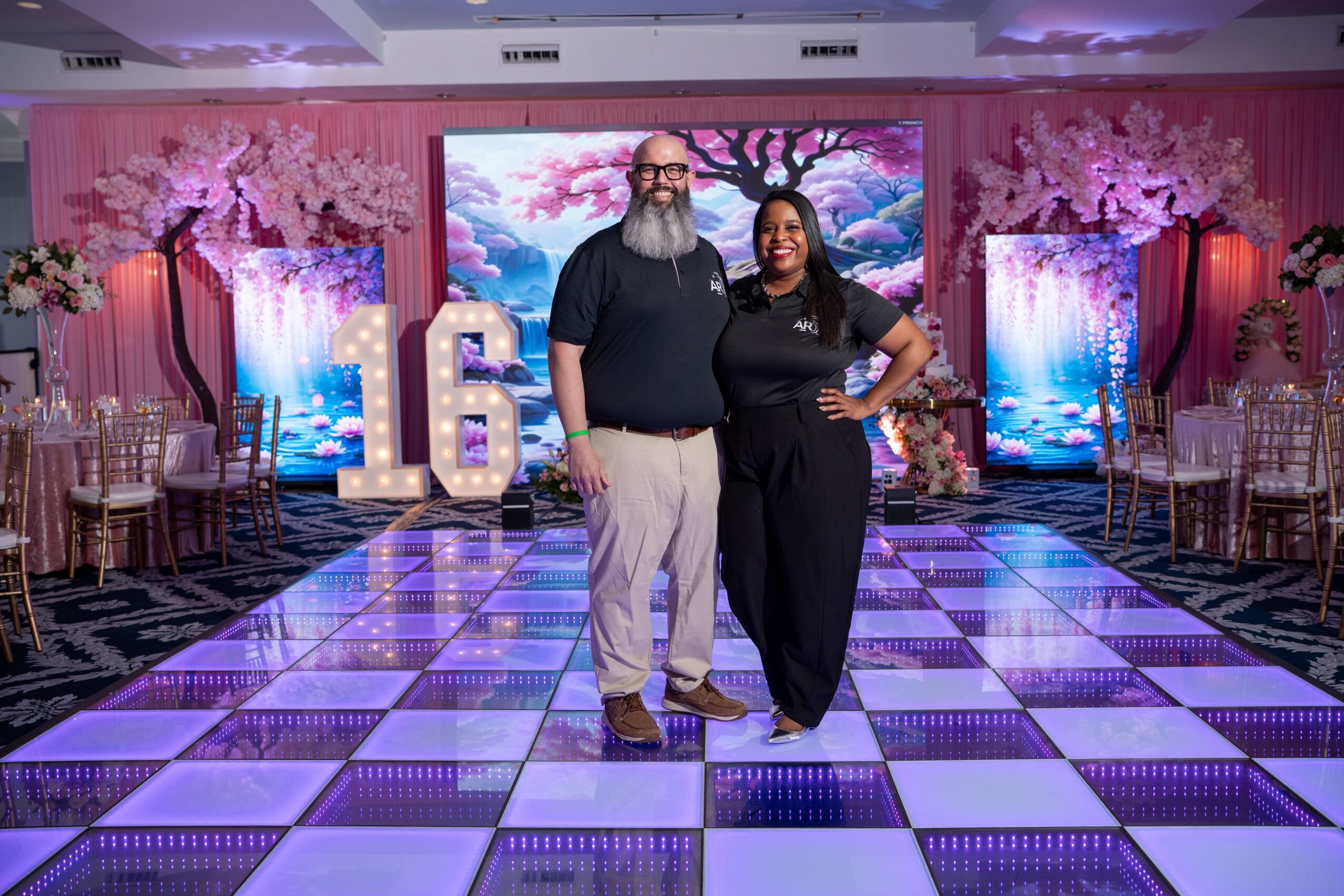 Two people stand on a decorated event stage with pink and purple lighting, large numbers '16', and digital screens in the background.