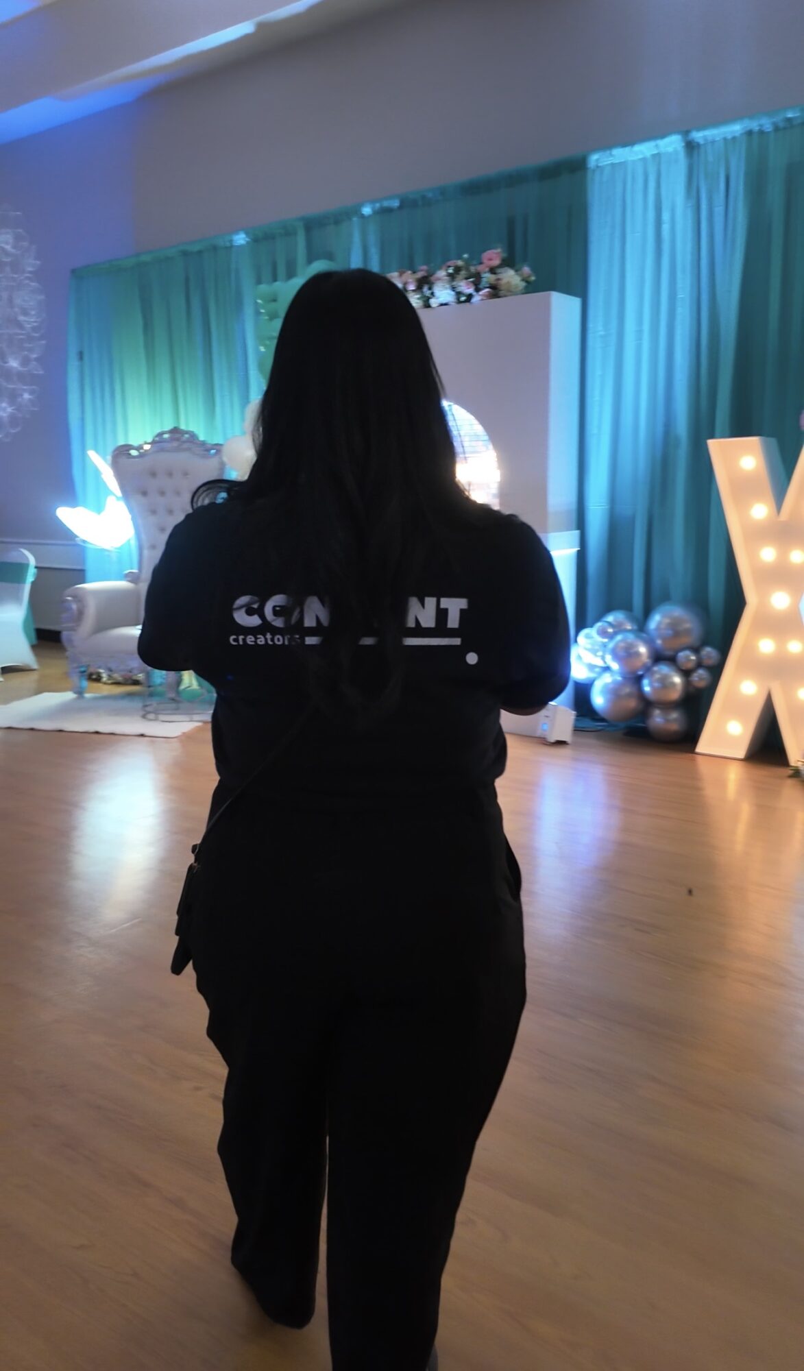 Person with long dark hair facing a decorated stage with teal curtains and large illuminated letters, indoors.