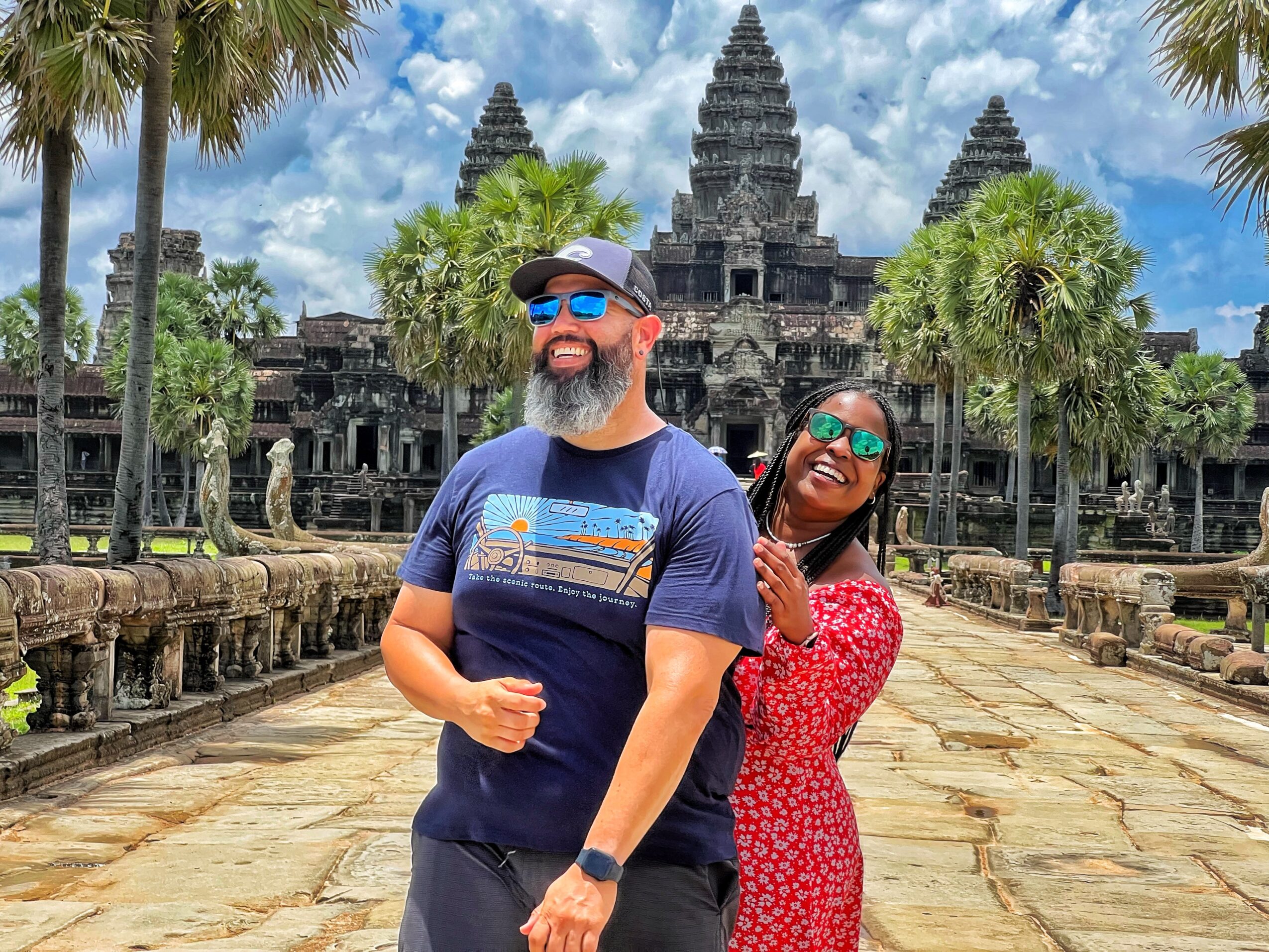 Two people smiling in front of an ancient temple with palm trees and a cloudy sky.