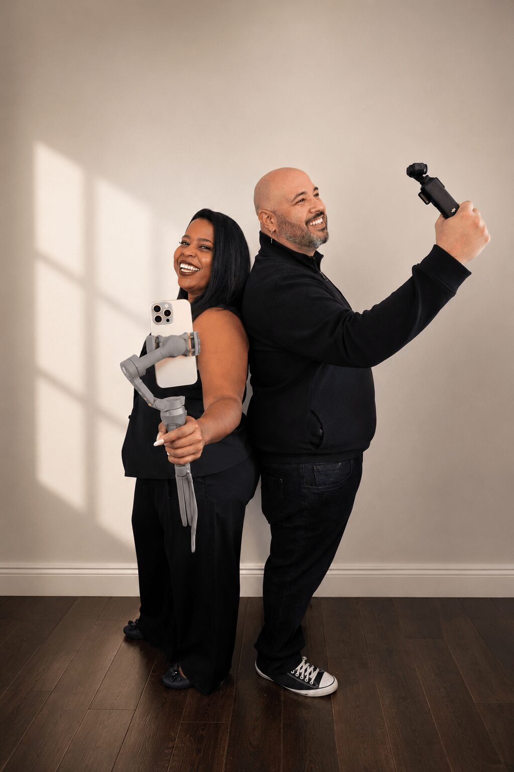 Two people stand back-to-back holding tools, smiling, in a room with a plain wall and window shadows.