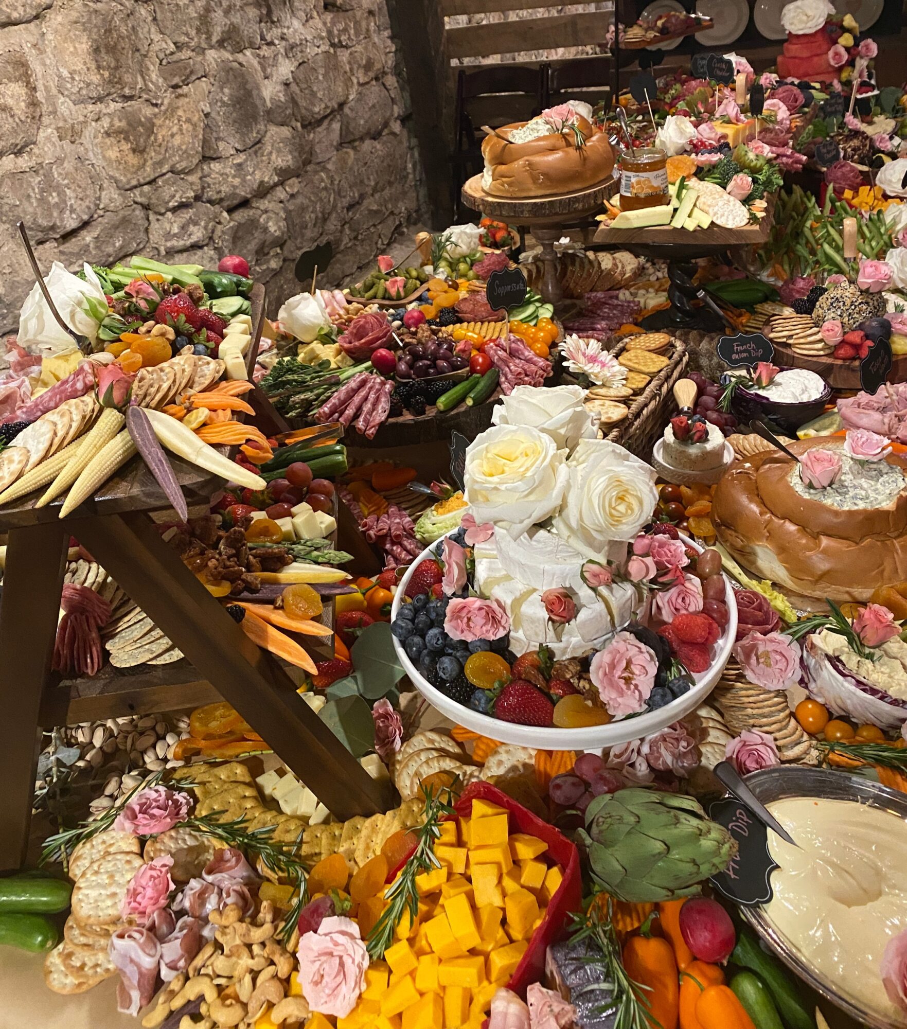 Table filled with cakes, fruits, flowers, and desserts, with a stone wall in the background.