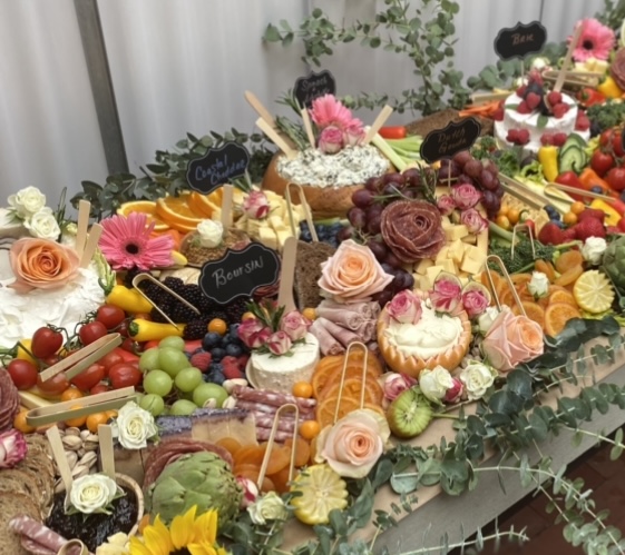 Table with various desserts, flowers, and fruits, decorated with greenery and small chalkboard signs, in a bright setting.