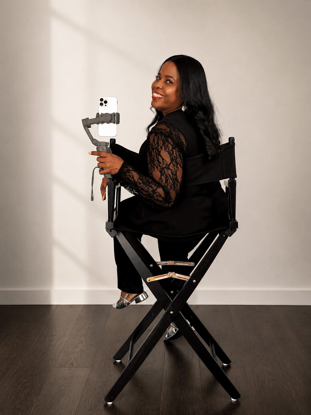 Woman sitting on a chair holding a camera, smiling, with a plain background.