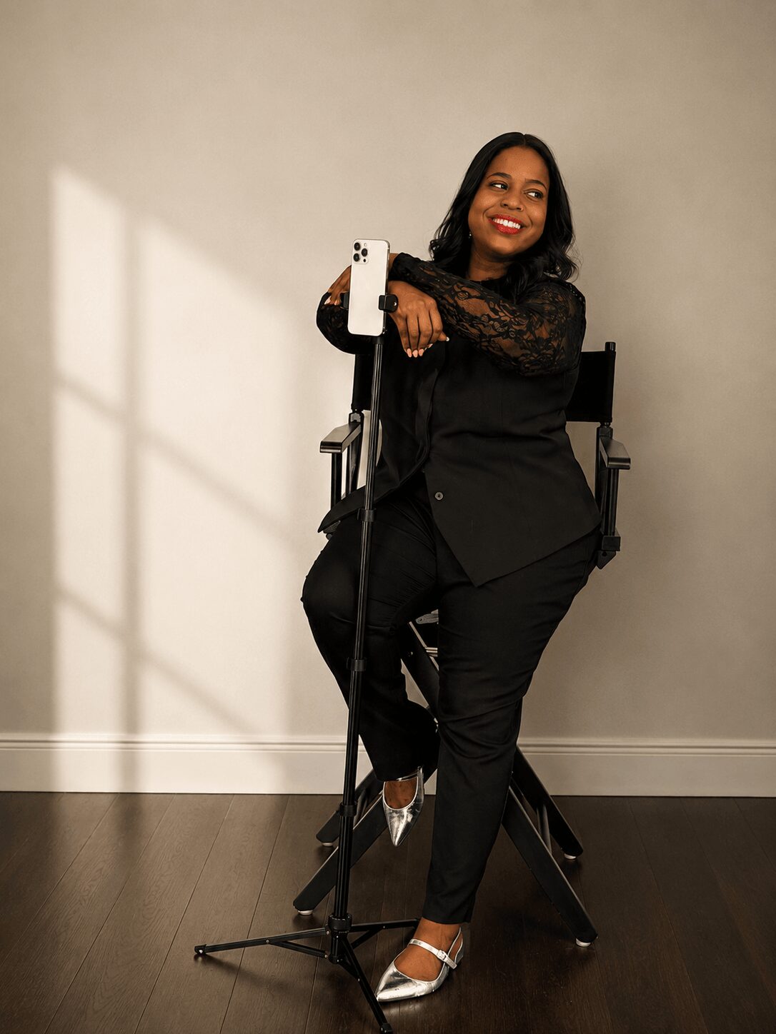 Woman sitting on a chair, taking a selfie with a smartphone, smiling, wearing black lace top and black pants, in a room with plain wall.