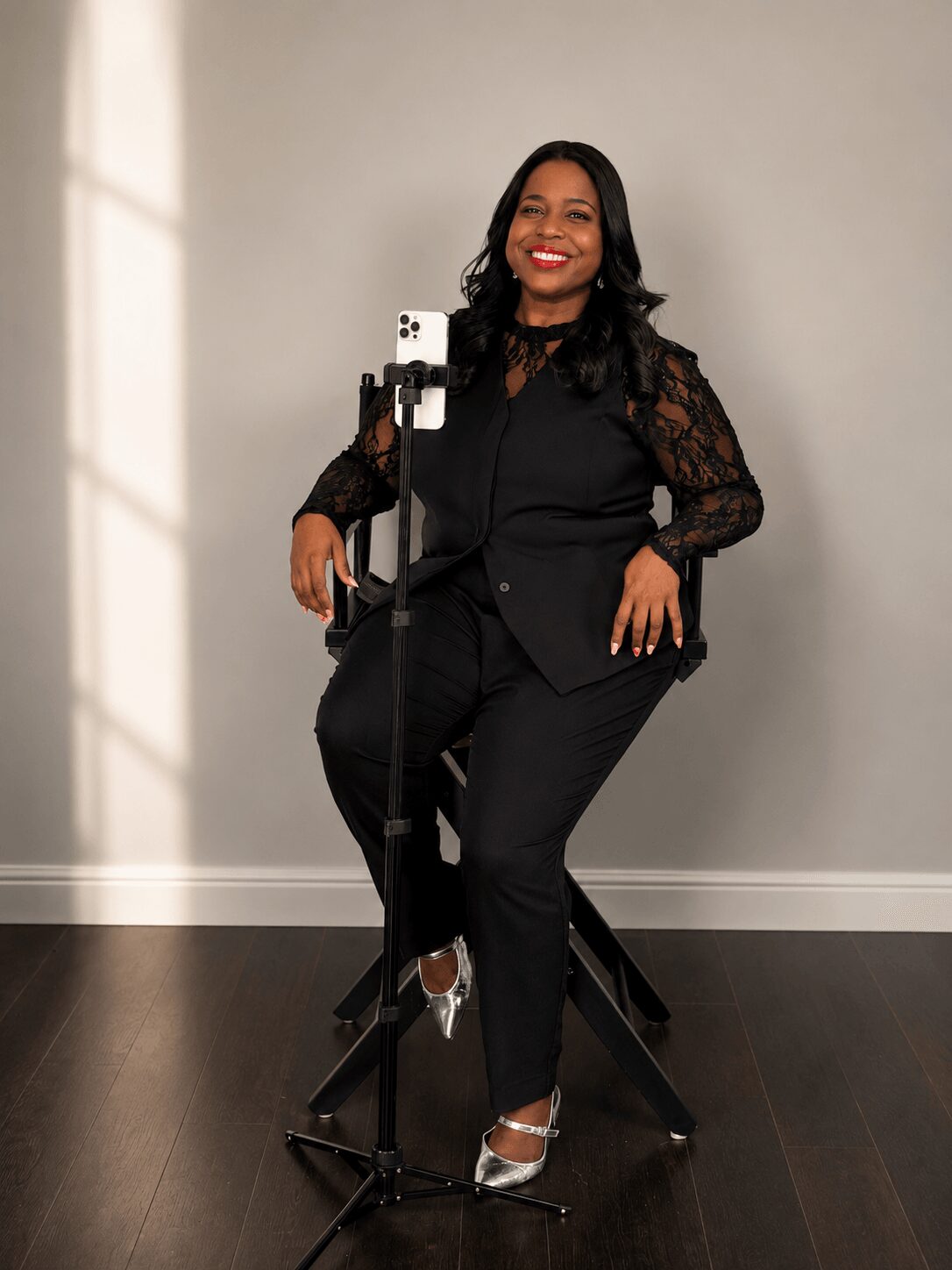 Woman sitting on a chair, smiling, wearing black outfit with lace sleeves, in a room with a plain wall and window shadow.