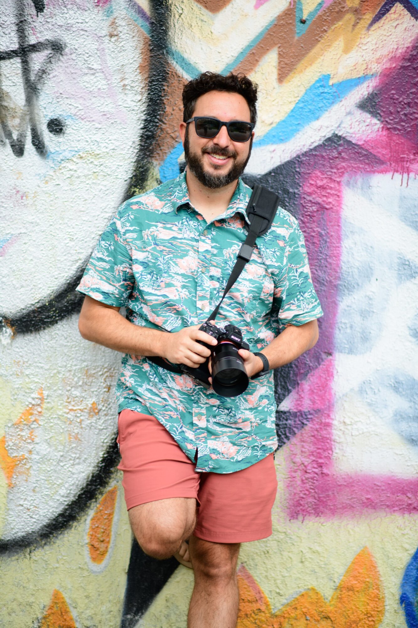 Man with sunglasses holding a camera, standing against a colorful graffiti wall, smiling, wearing a patterned shirt and shorts.