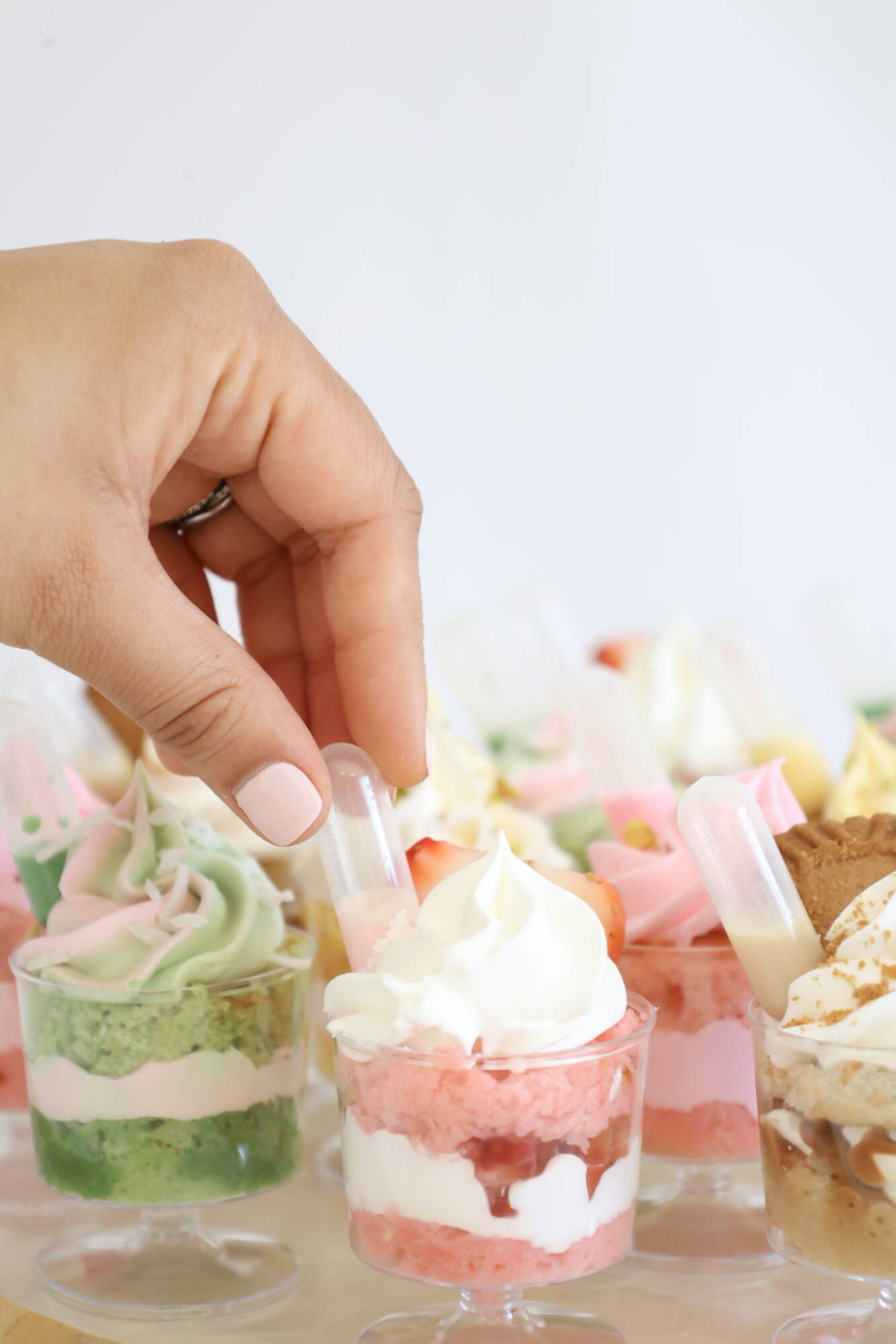 Hand reaching for a layered dessert in a small glass with pink and white swirls, surrounded by other desserts, against a light background.