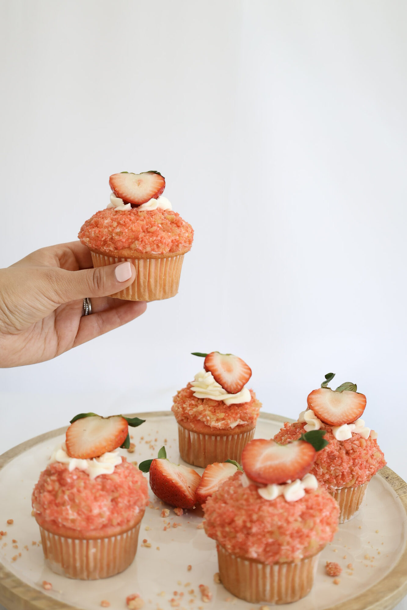 Hand holding a cupcake with strawberry topping, other cupcakes on a plate with strawberries, white background