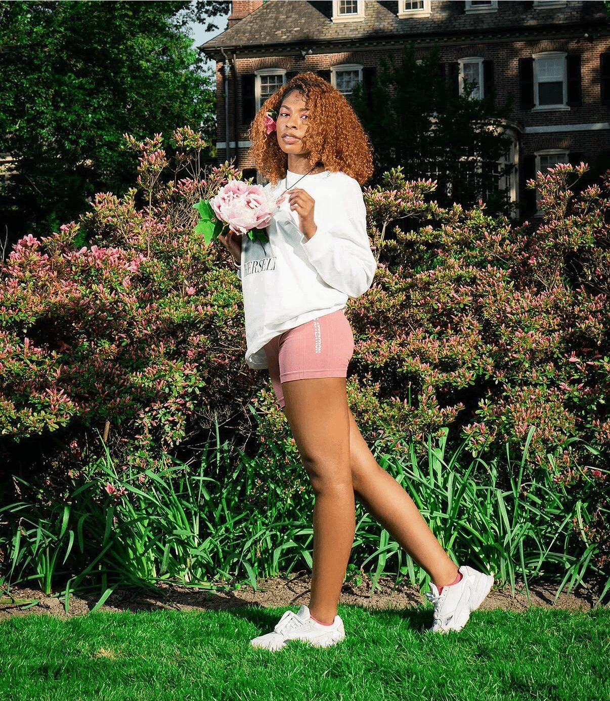 Young woman with curly hair holding flowers in a garden with blooming bushes and a house in the background.