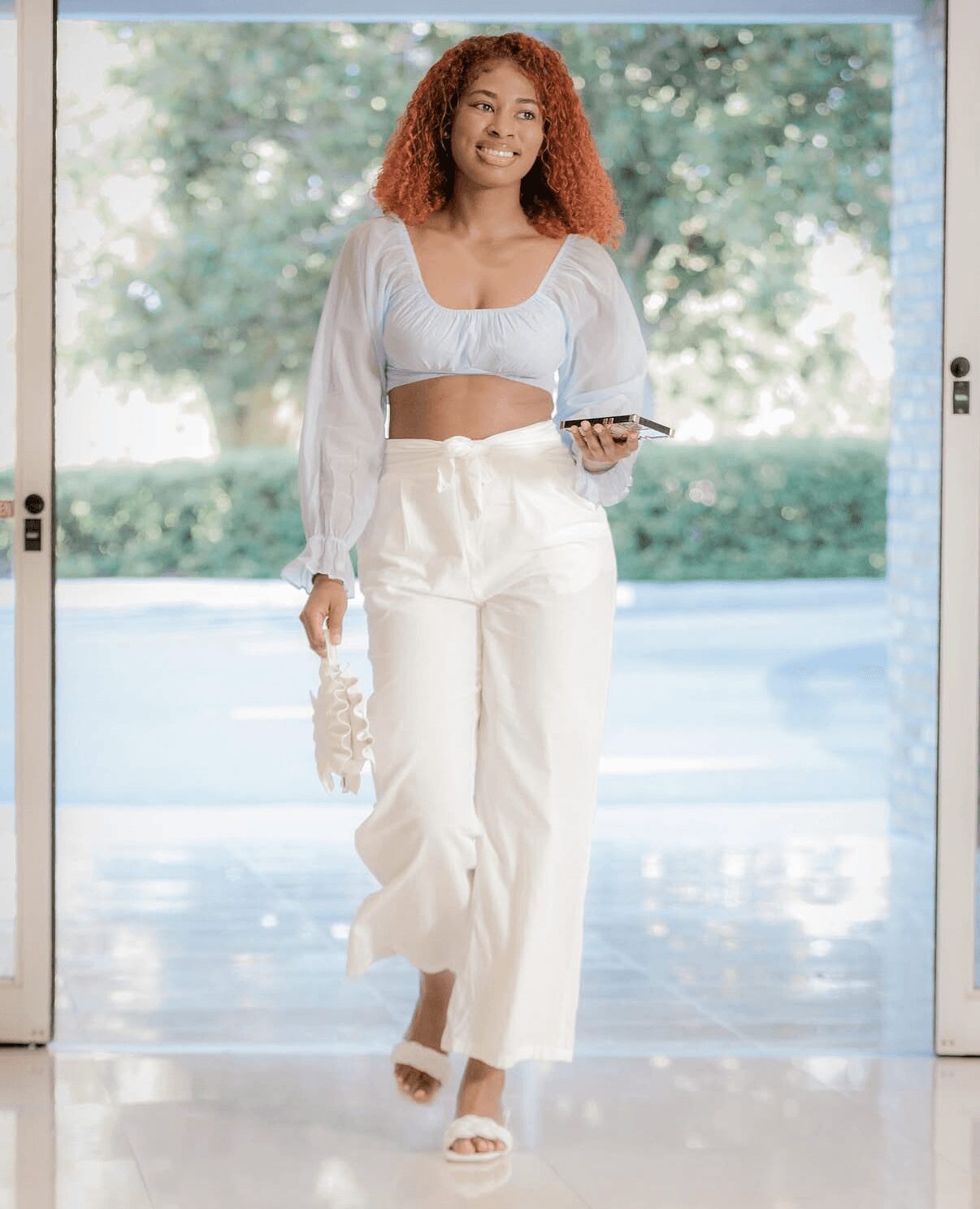Woman with curly hair walking outdoors holding a phone and a bag, wearing a white crop top and pants, sunny day.