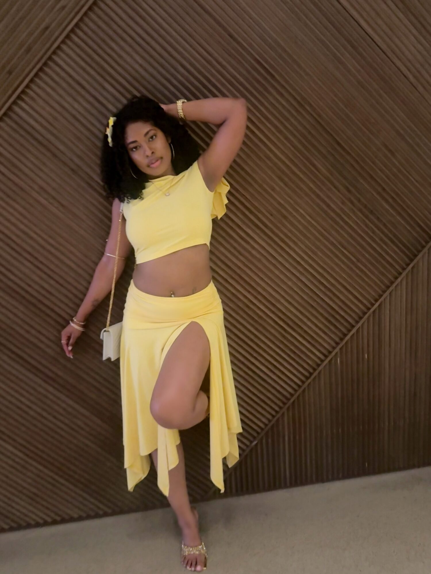 Young woman in yellow crop top and skirt poses with one leg raised against wooden wall.