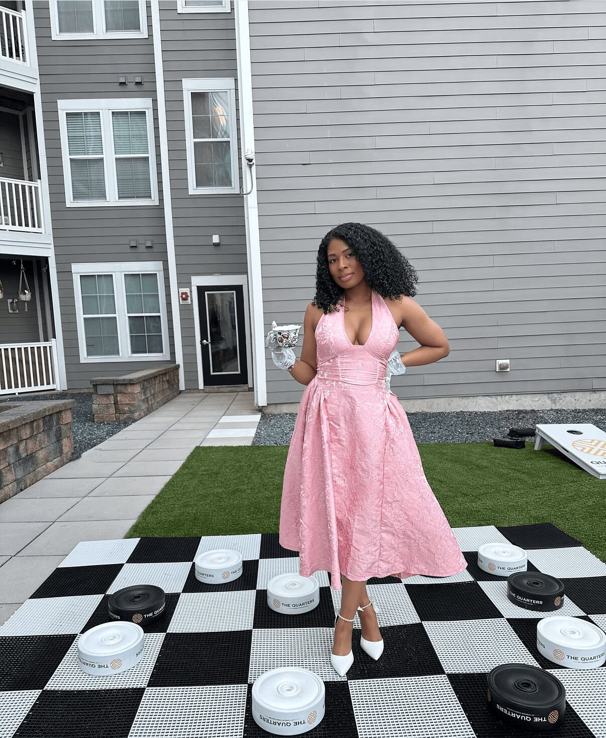 Woman in pink dress standing on a checkered outdoor game board with white and black pieces, in front of a residential building.