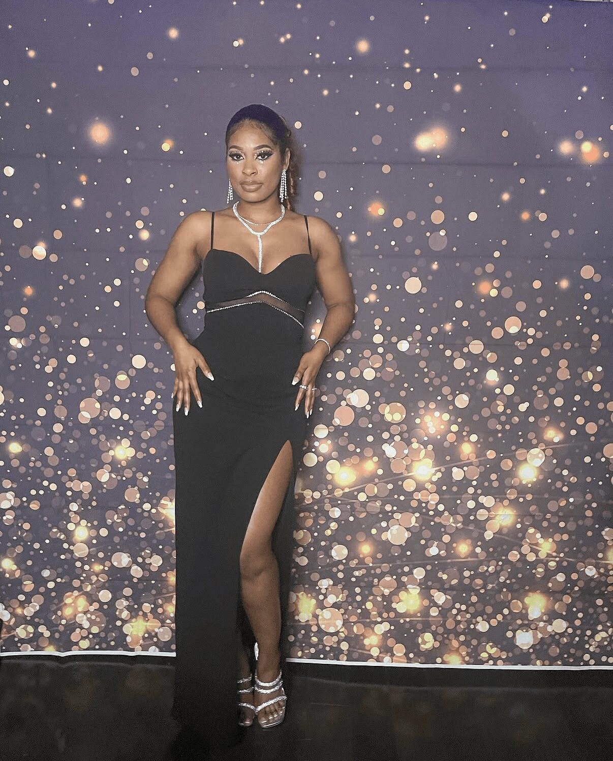Woman in black dress with slit, standing against a starry background, wearing jewelry and high heels.