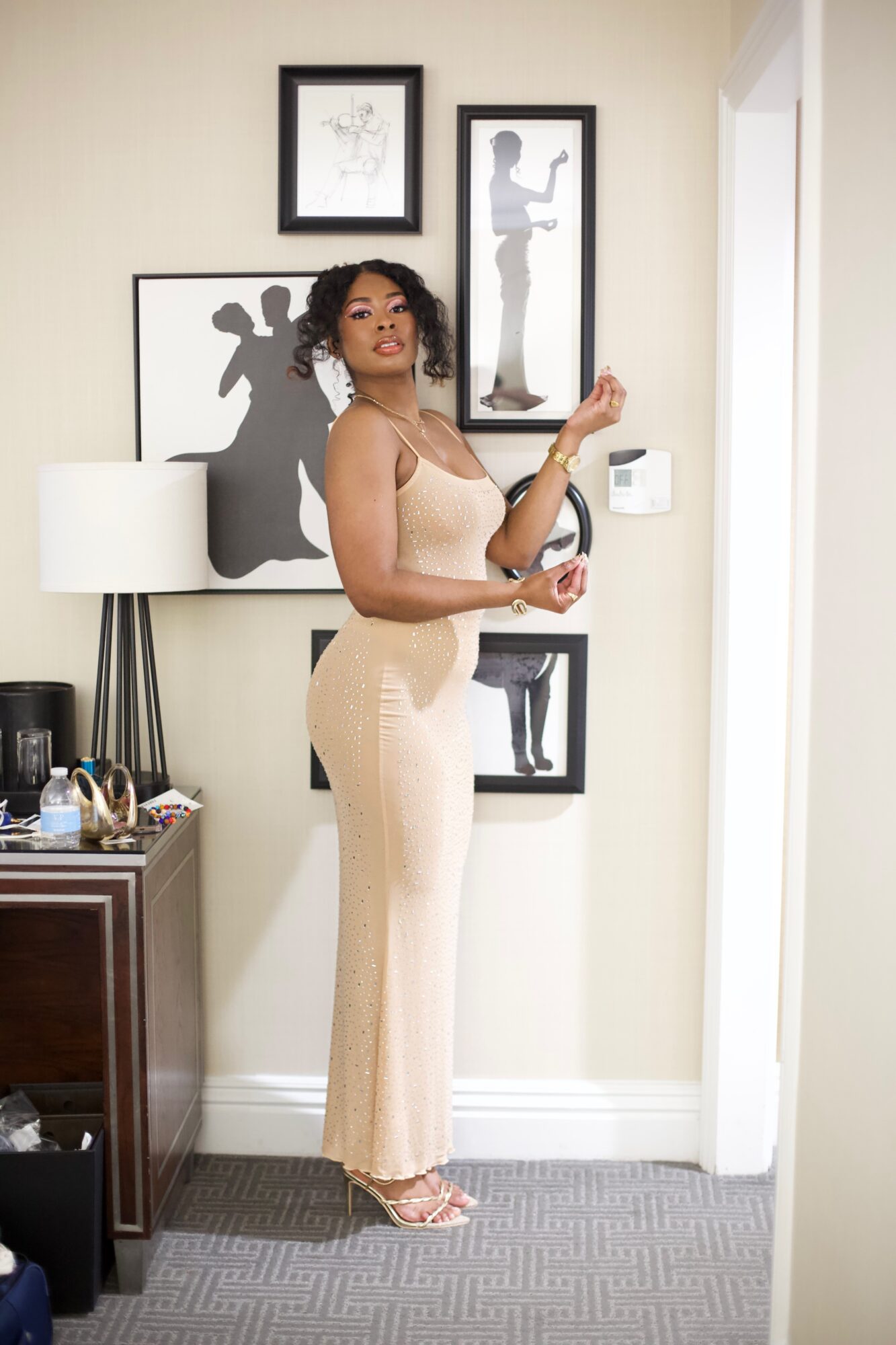 Woman standing in a room with framed black and white artwork on the wall, holding a glass, wearing beige pants and a top.