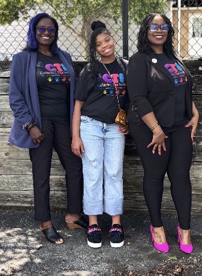 Three women standing outdoors in front of a fence, smiling, wearing black T-shirts with colorful designs, and casual pants.