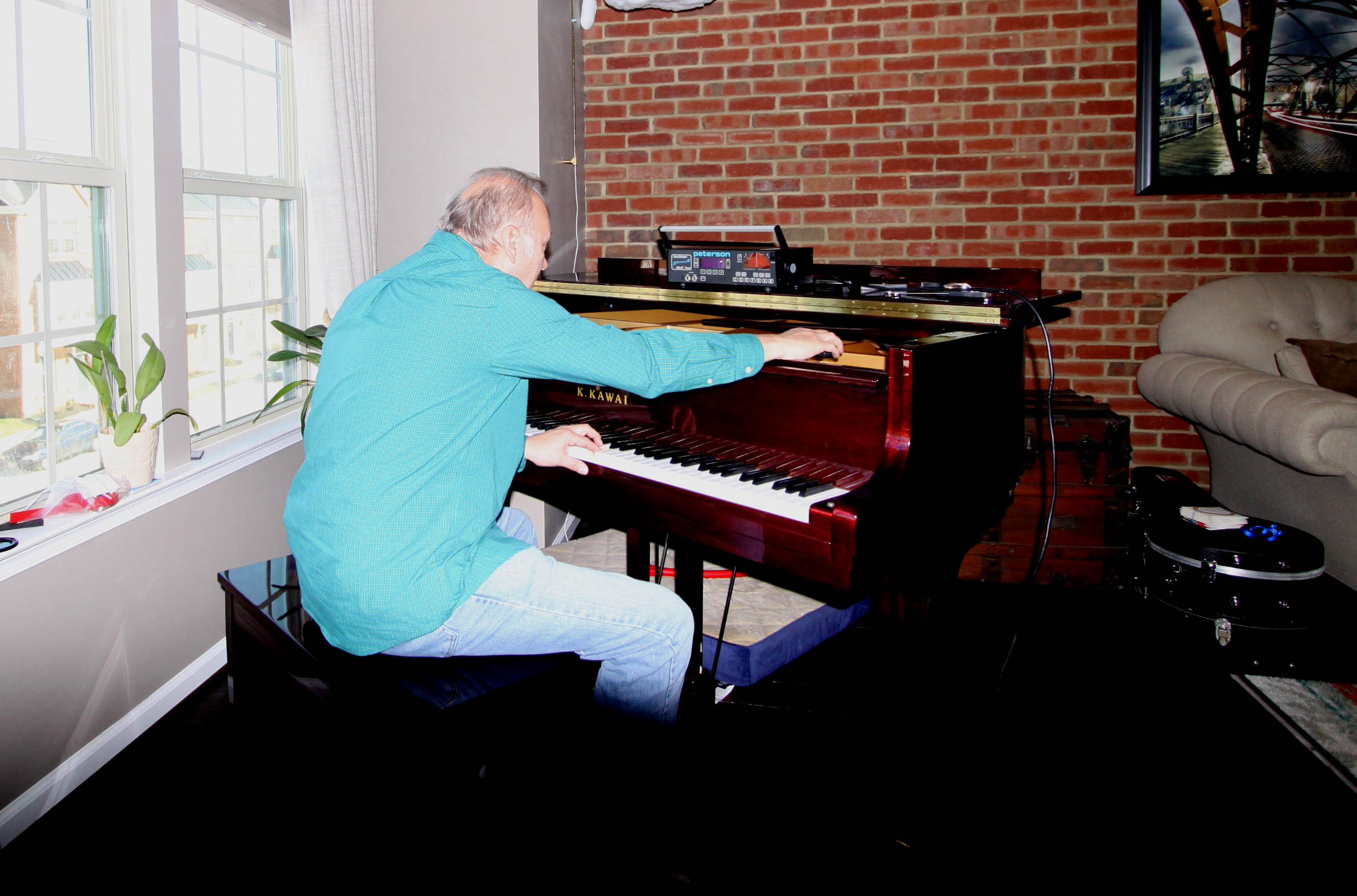 Person playing a grand piano in a room with brick wall and large window, sitting on a black bench.