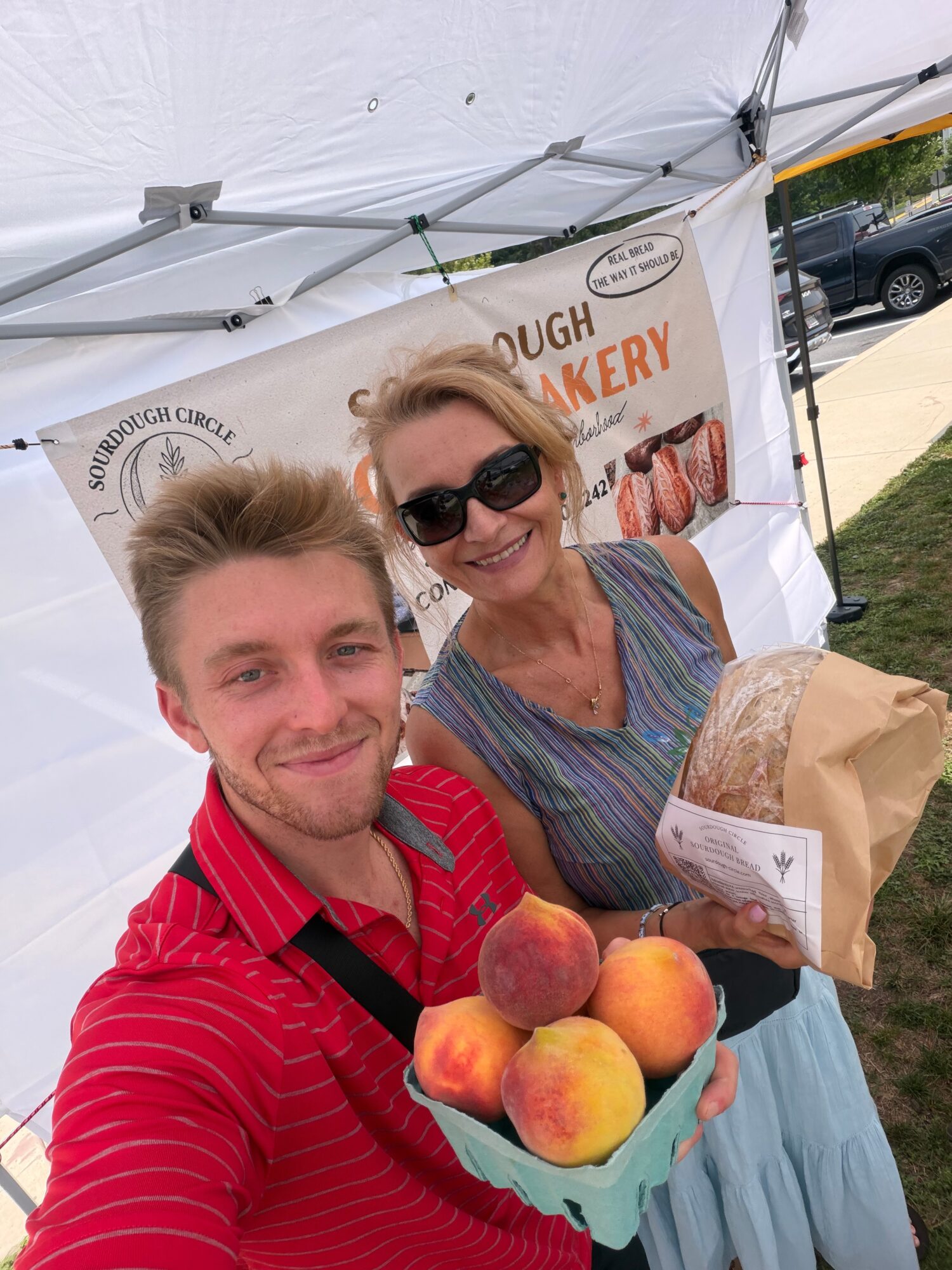 A young man and an older woman smiling at the camera, holding peaches and a paper bag, standing under a tent.