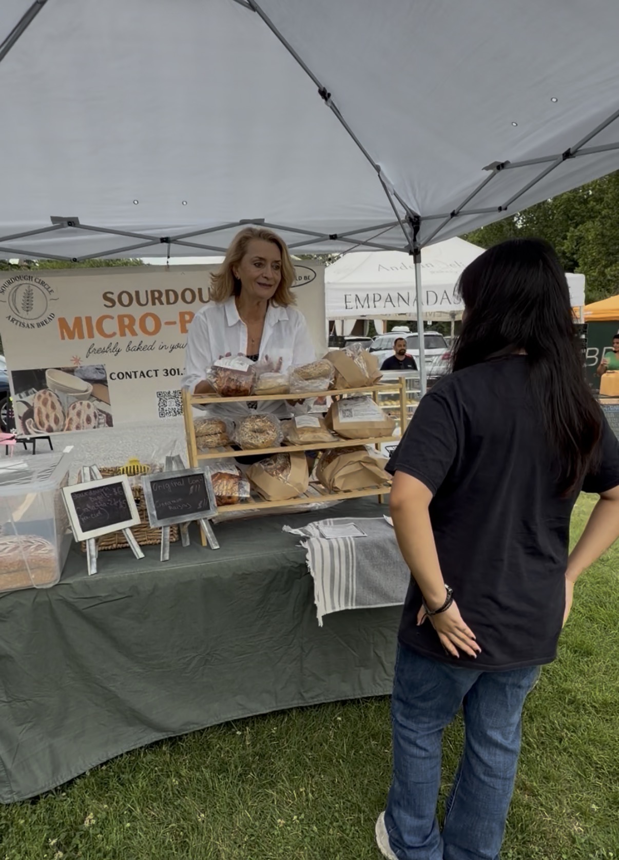 Woman behind a table at an outdoor market stall, talking to a customer, with bread and signs displayed, under a canopy.