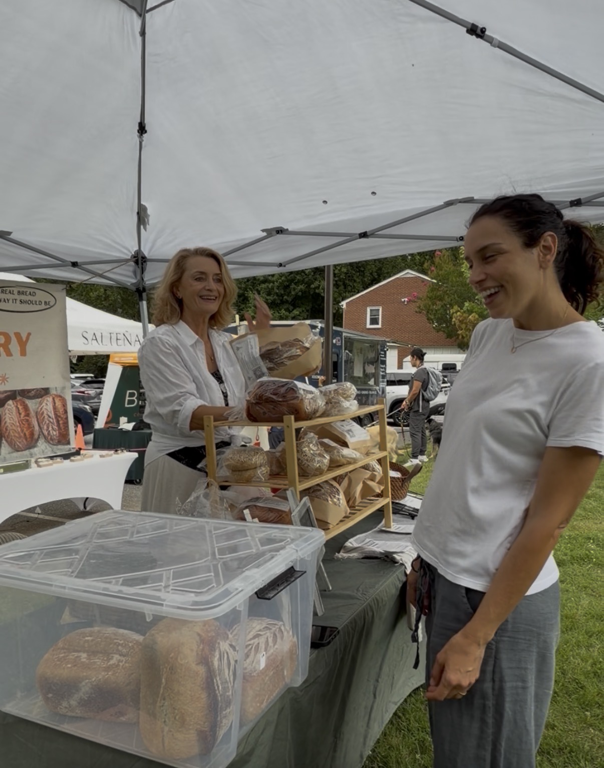Two women smiling at a market stall with baked goods under a white tent, outdoors on a cloudy day.