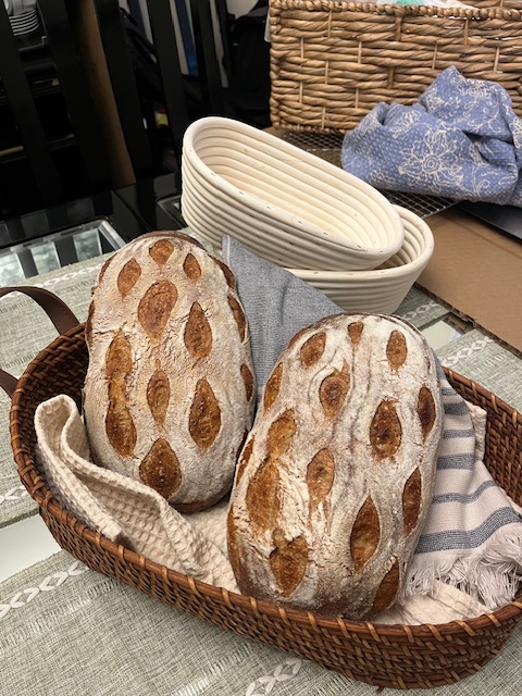 Two loaves of bread with a pattern of seeds on top in a wicker basket lined with cloth, placed on a table.