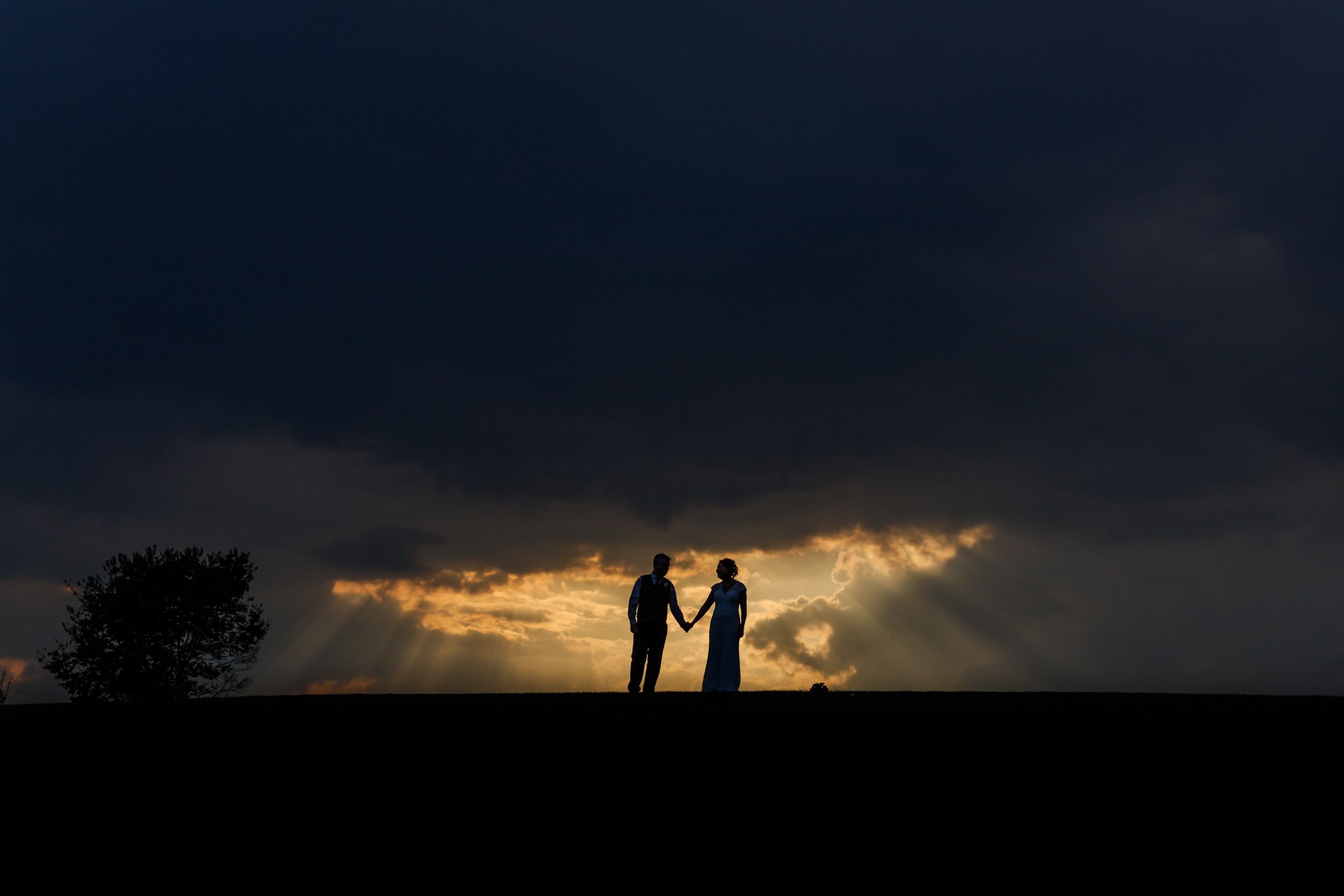 Two people holding hands under a dark, cloudy sky with sunlight breaking through, silhouetted against the horizon.