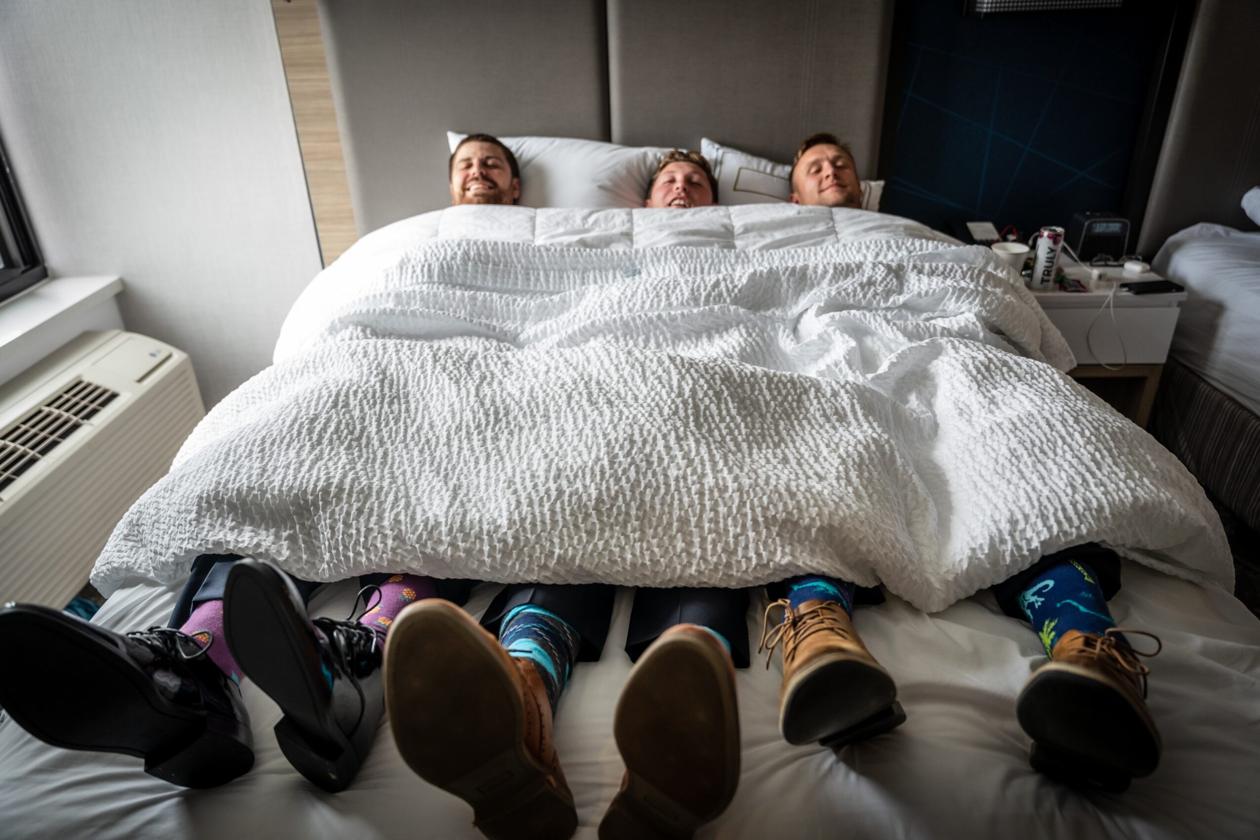 Three people lying in bed with only their feet visible, wearing colorful socks and shoes, in a hotel room.