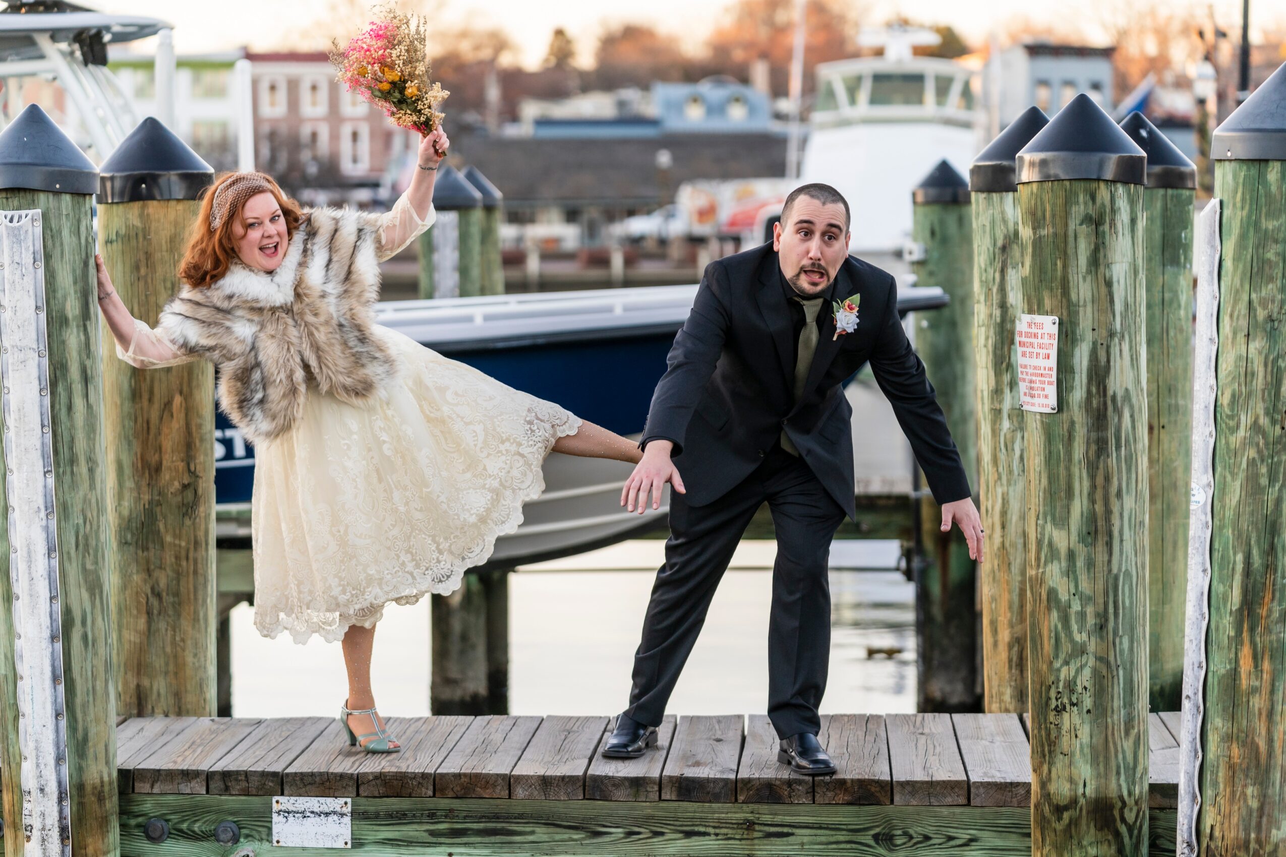 A woman in a wedding dress and fur coat, holding a bouquet, is leaning on a man in a suit on a dock. She is smiling and holding a bouquet, while he is crouching with a surprised expression. Wooden posts and water are in the background.