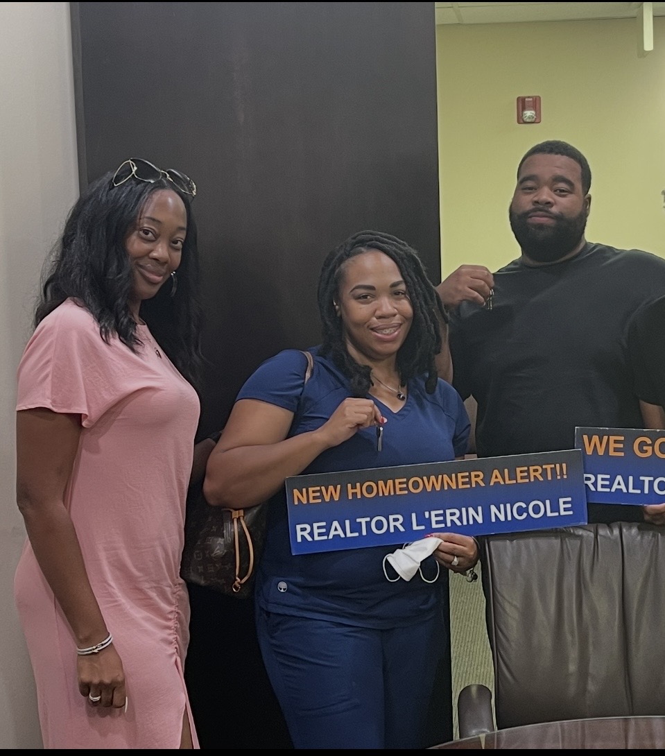 Three people standing indoors, one holding a sign that reads 'Realtor L'erin Nicole' and 'New Homeowner Alert'.