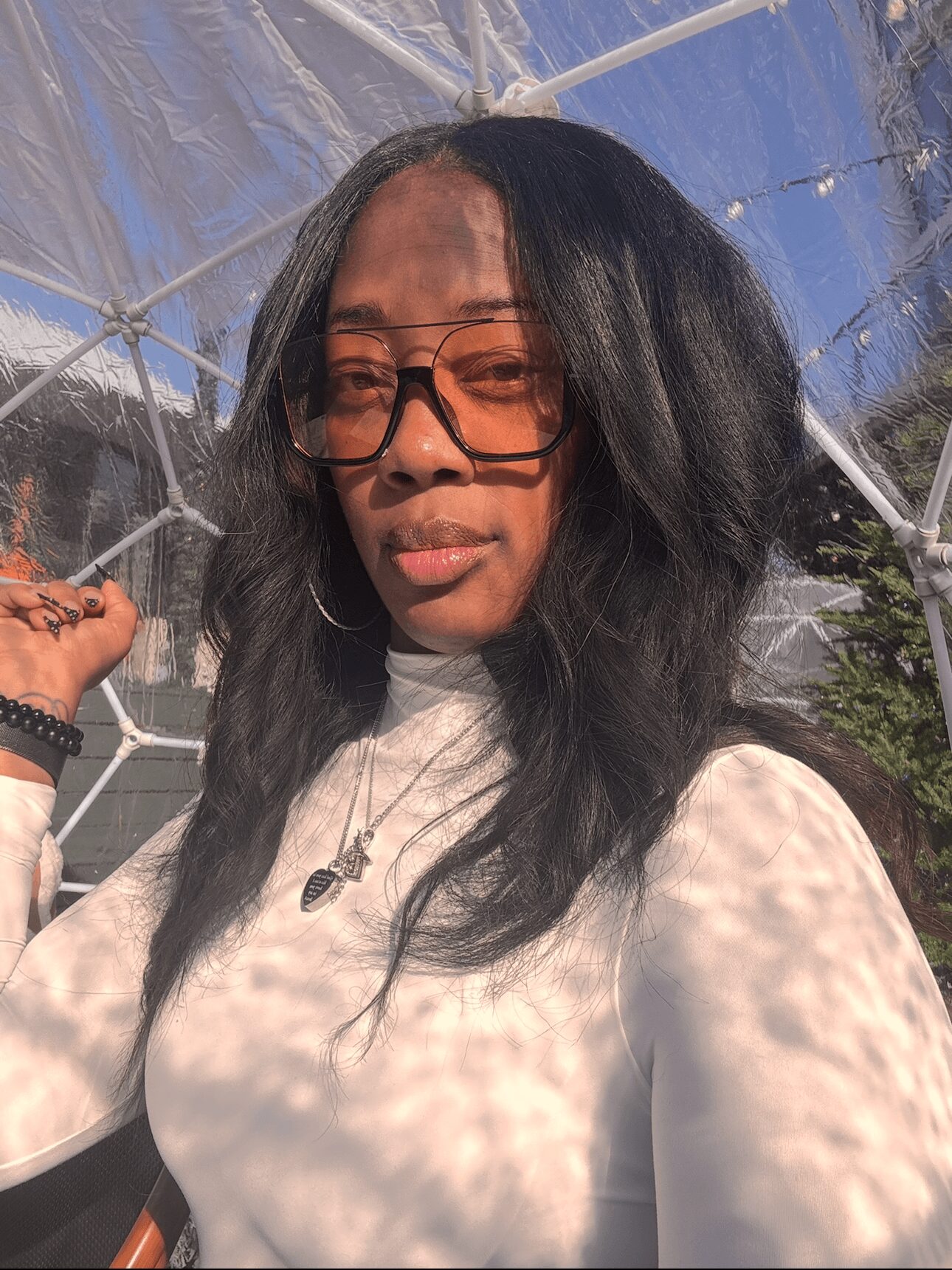 Woman with long dark hair, glasses, and a white top, standing outdoors with sunlight and shadows on her face.