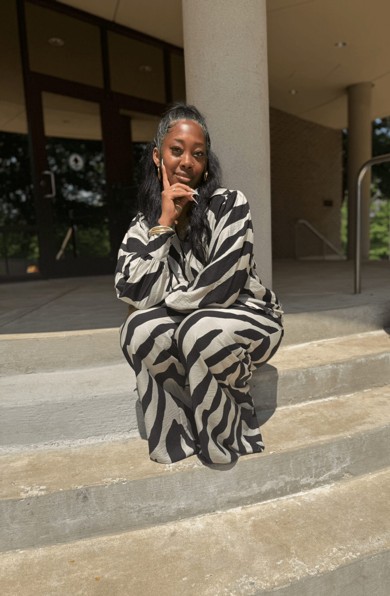 Person sitting on steps outside, wearing a zebra-patterned outfit, with hand near face, smiling.