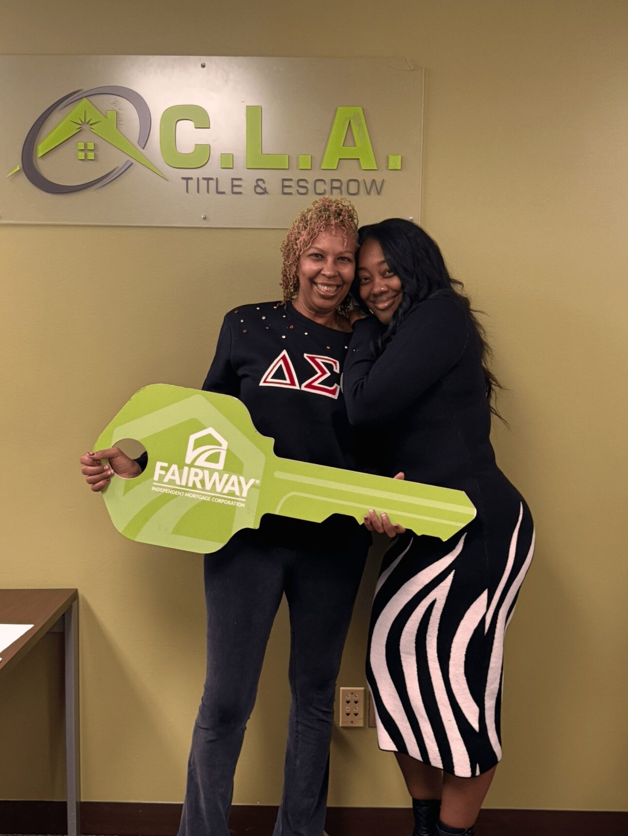 Two women smiling and hugging, one holding a large green key with 'Fairway' logo, standing in front of a sign reading 'C.L.A. Title & Escrow'.