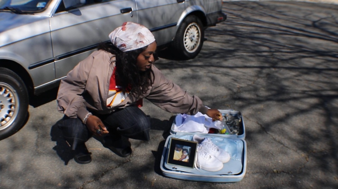 Woman crouching on street, looking into an open suitcase with various items, parked car in background.