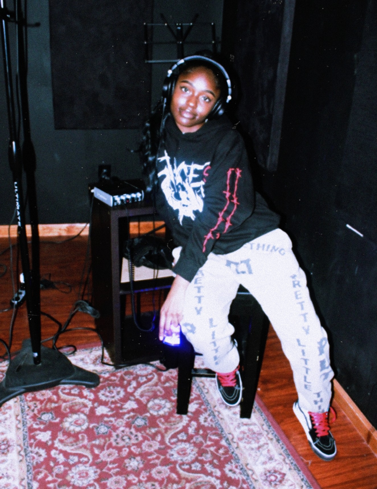 Child sitting on a chair with headphones, wearing a black hoodie and white pants, in a recording studio. Word on pants: 'LITTLE'.
