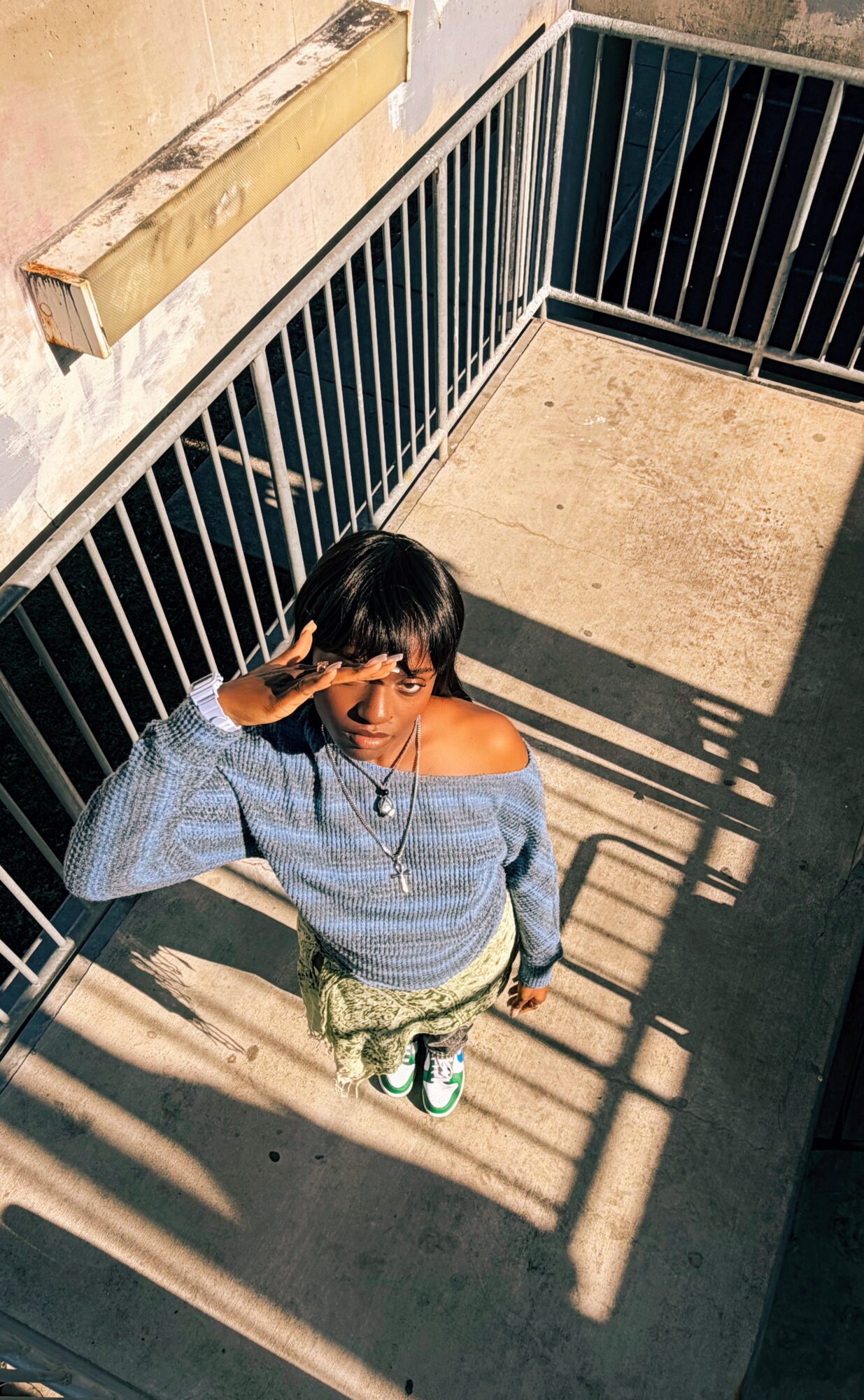 Young woman with dark hair, wearing a blue off-shoulder sweater and patterned pants, standing on a balcony with shadows.