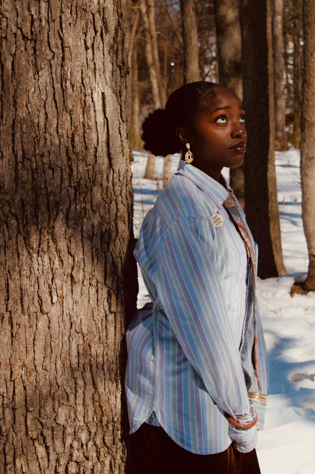 Woman with dark skin and curly hair stands in a snowy forest, leaning against a tree, wearing a striped shirt.