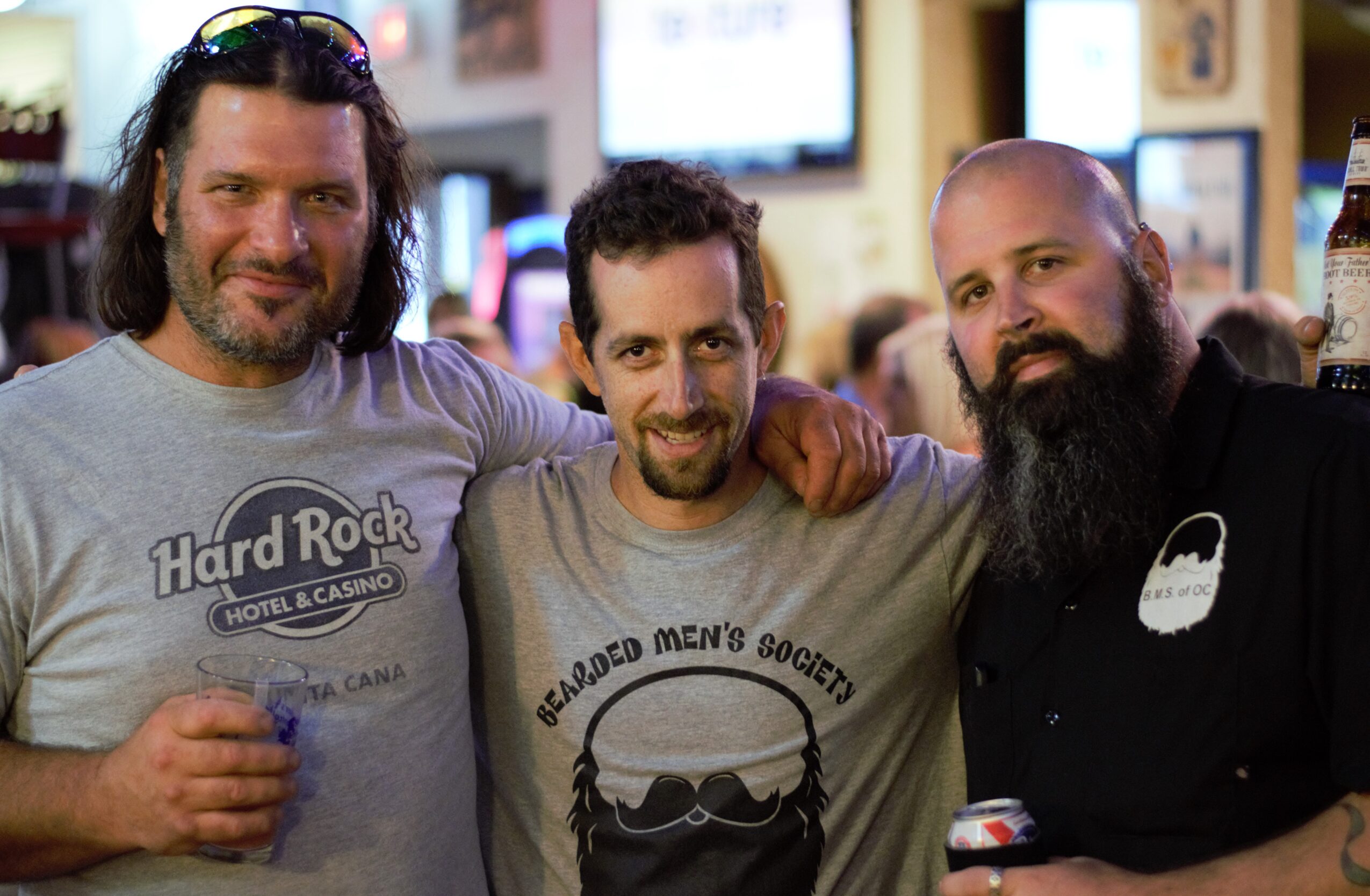 Three men with beards and casual clothing pose together indoors, smiling at the camera.