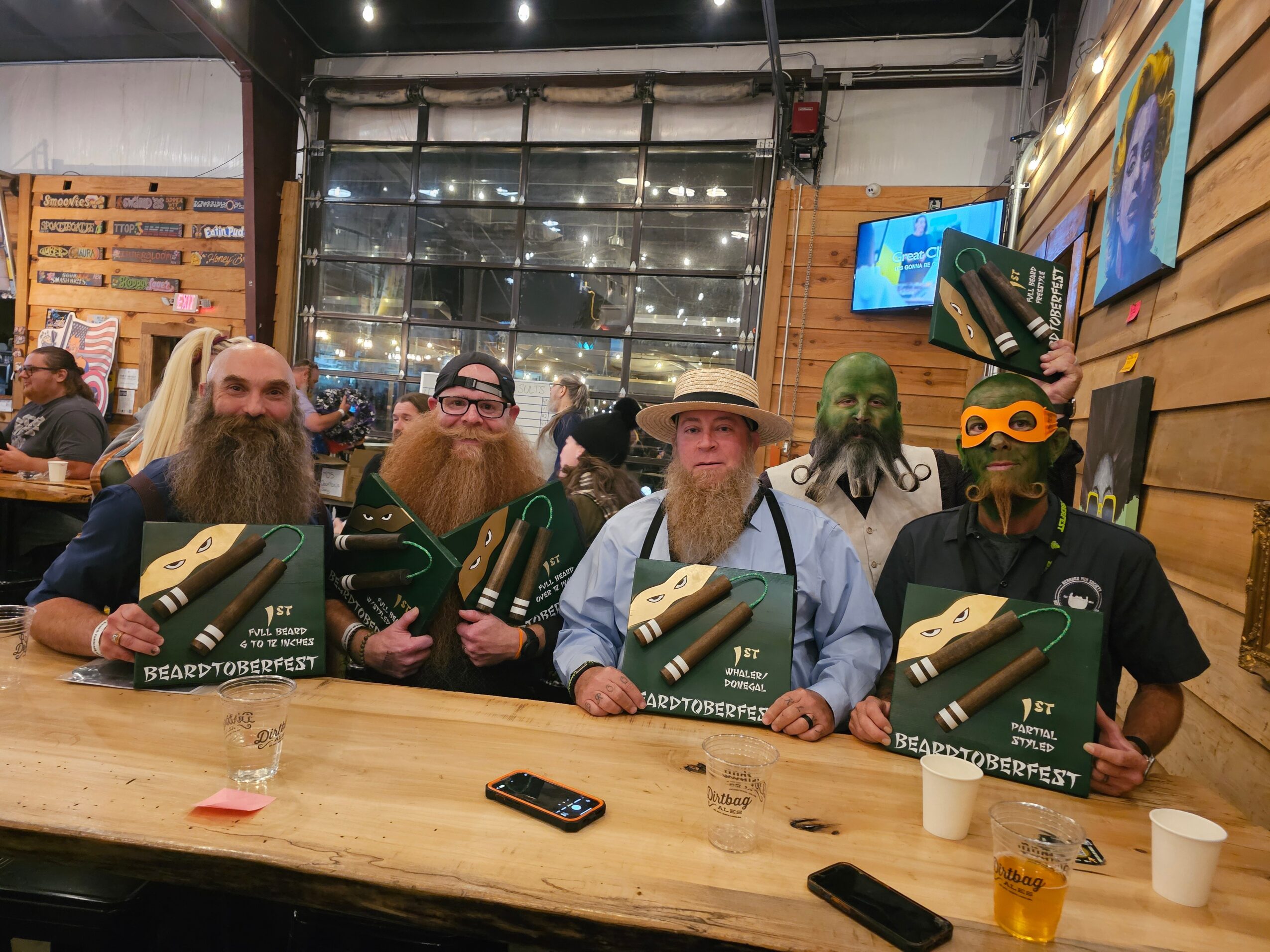 Group of five people with beards and masks holding boxes at a wooden table in a store, with screens and wooden walls behind them.