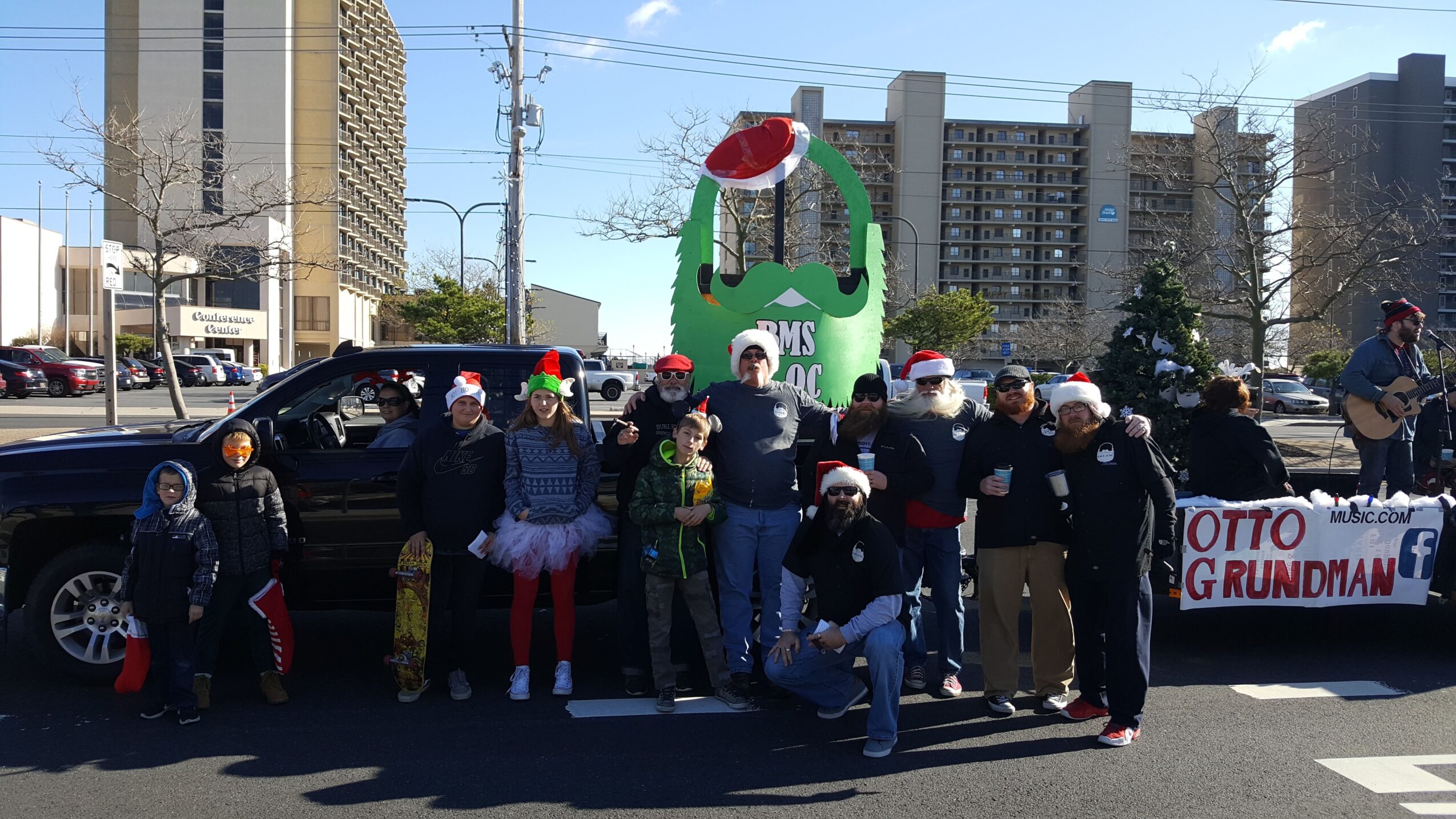 Group of people wearing Santa hats and masks standing in front of a decorated float and a black vehicle during a parade.