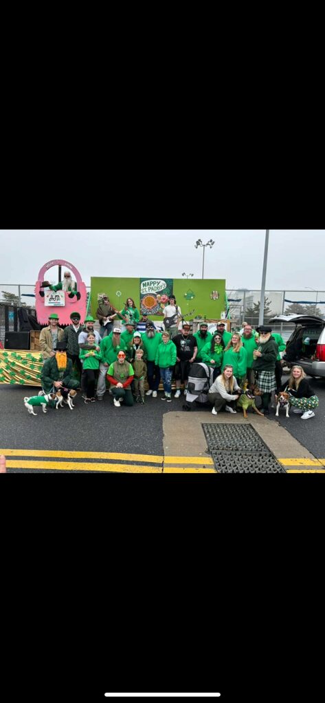 Group of people wearing green shirts standing and sitting in front of a food truck and a green backdrop, outdoors.