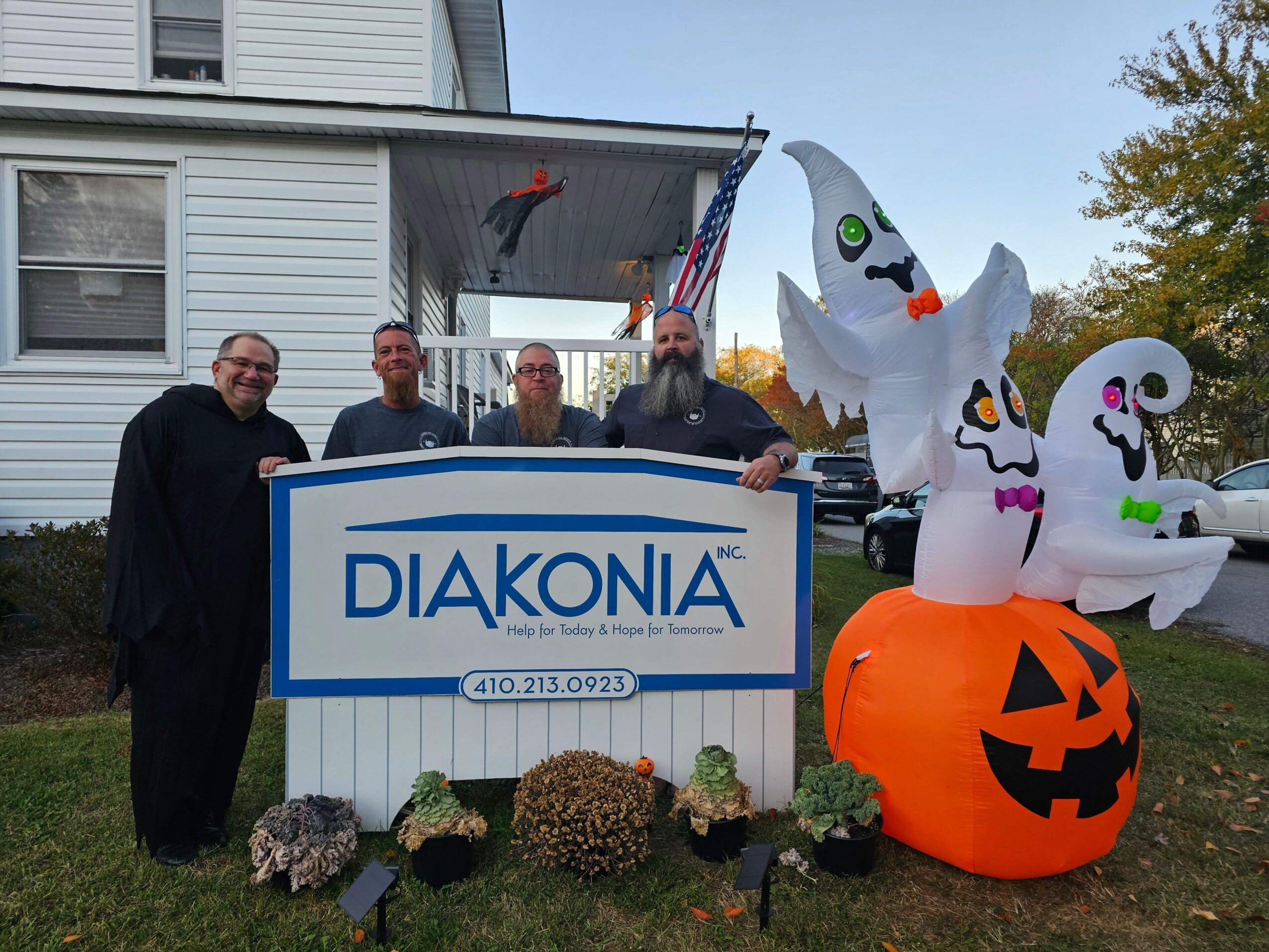 Four people stand behind a sign with Halloween decorations, including inflatable ghosts and a pumpkin, outside a house with a porch.