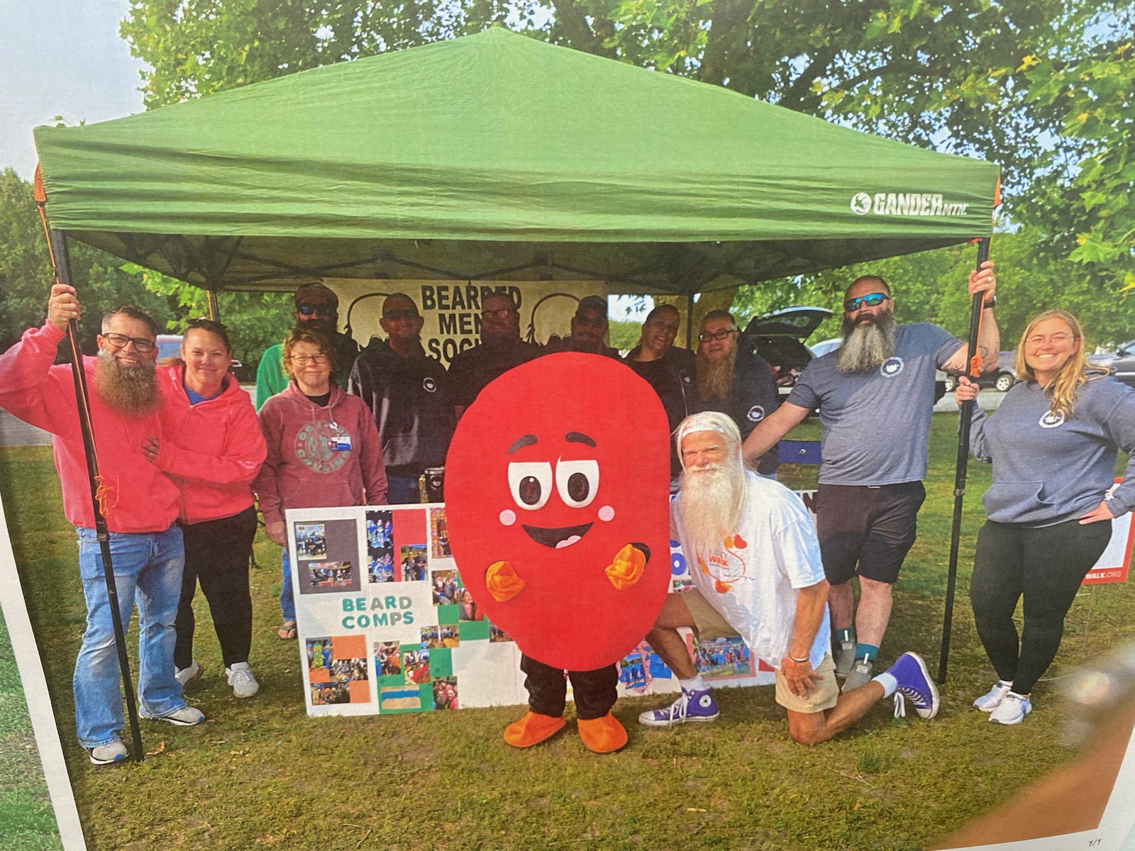Group of people outdoors under a green tent, including a person in a red mascot costume, some holding signs, and a person dressed as Santa Claus.