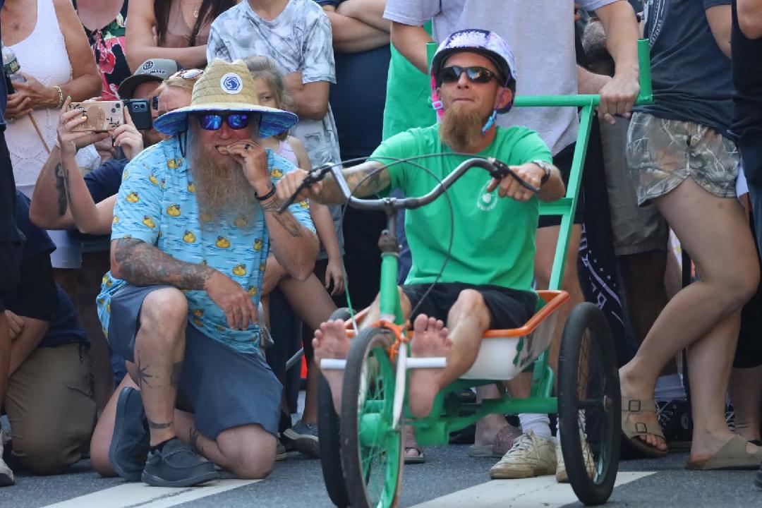 Child in a green shirt and sunglasses riding a tricycle surrounded by people at an outdoor event.
