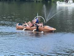People on an inflatable boat in water, with a person standing and spraying water, and a boat in the background