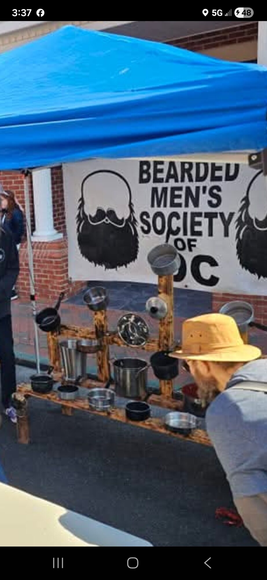 Person wearing a yellow hat browsing items at a market stall with a beard-themed sign in the background.