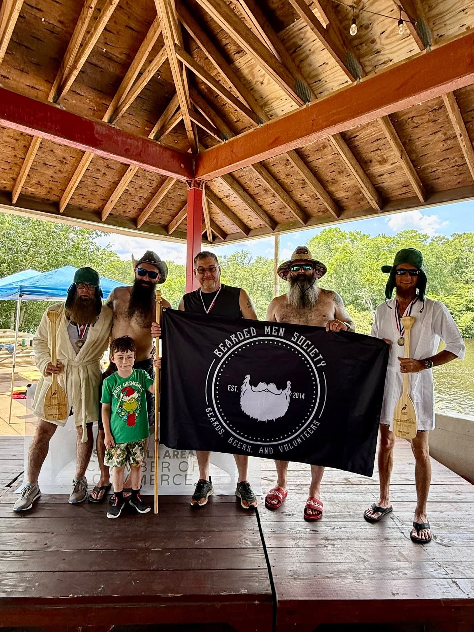 Group of people holding a black flag with a white logo, standing under a wooden pavilion outdoors.