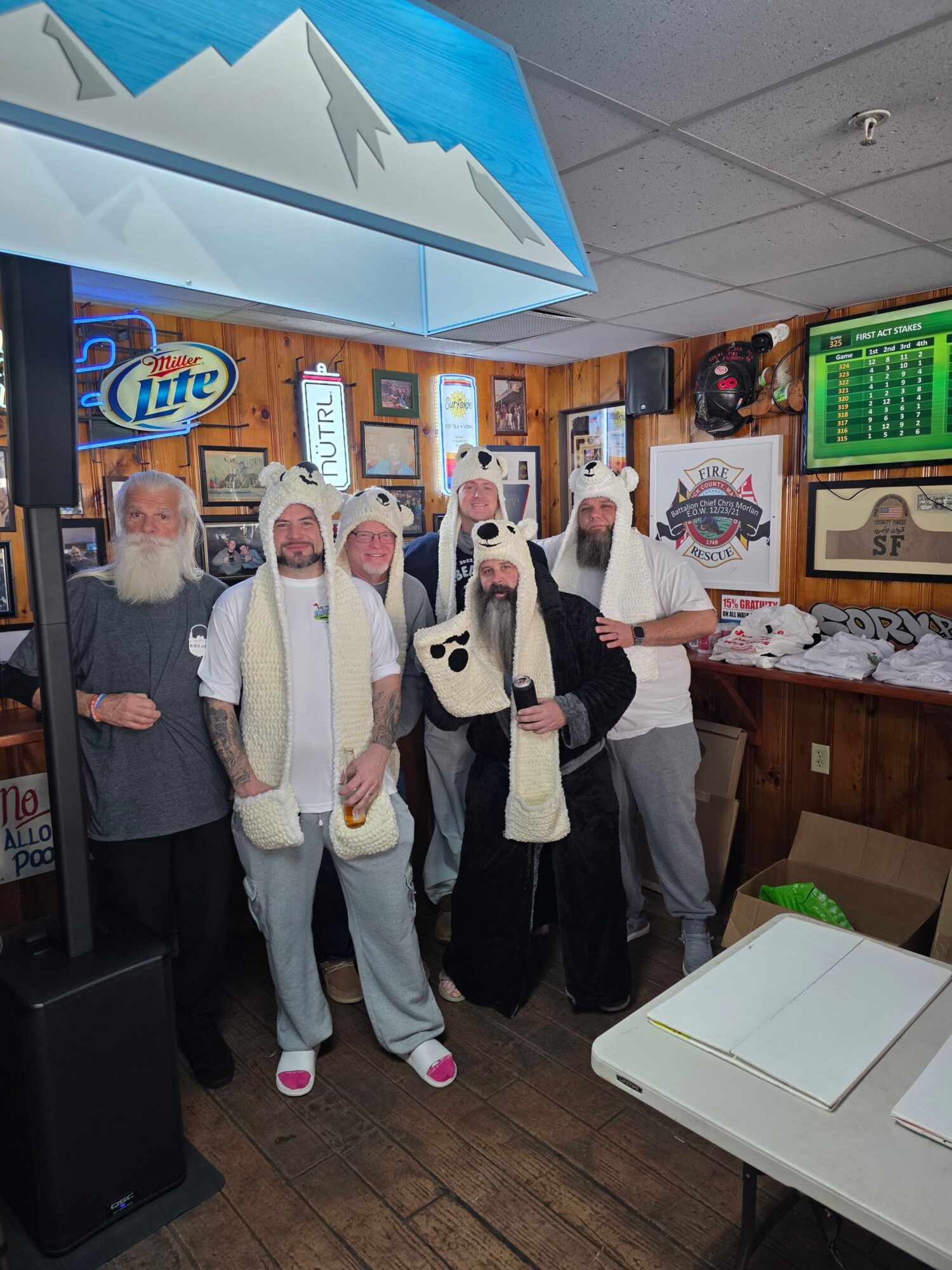 Four elderly men standing in a room, one holding a guitar, with framed pictures and a neon sign on the wall.